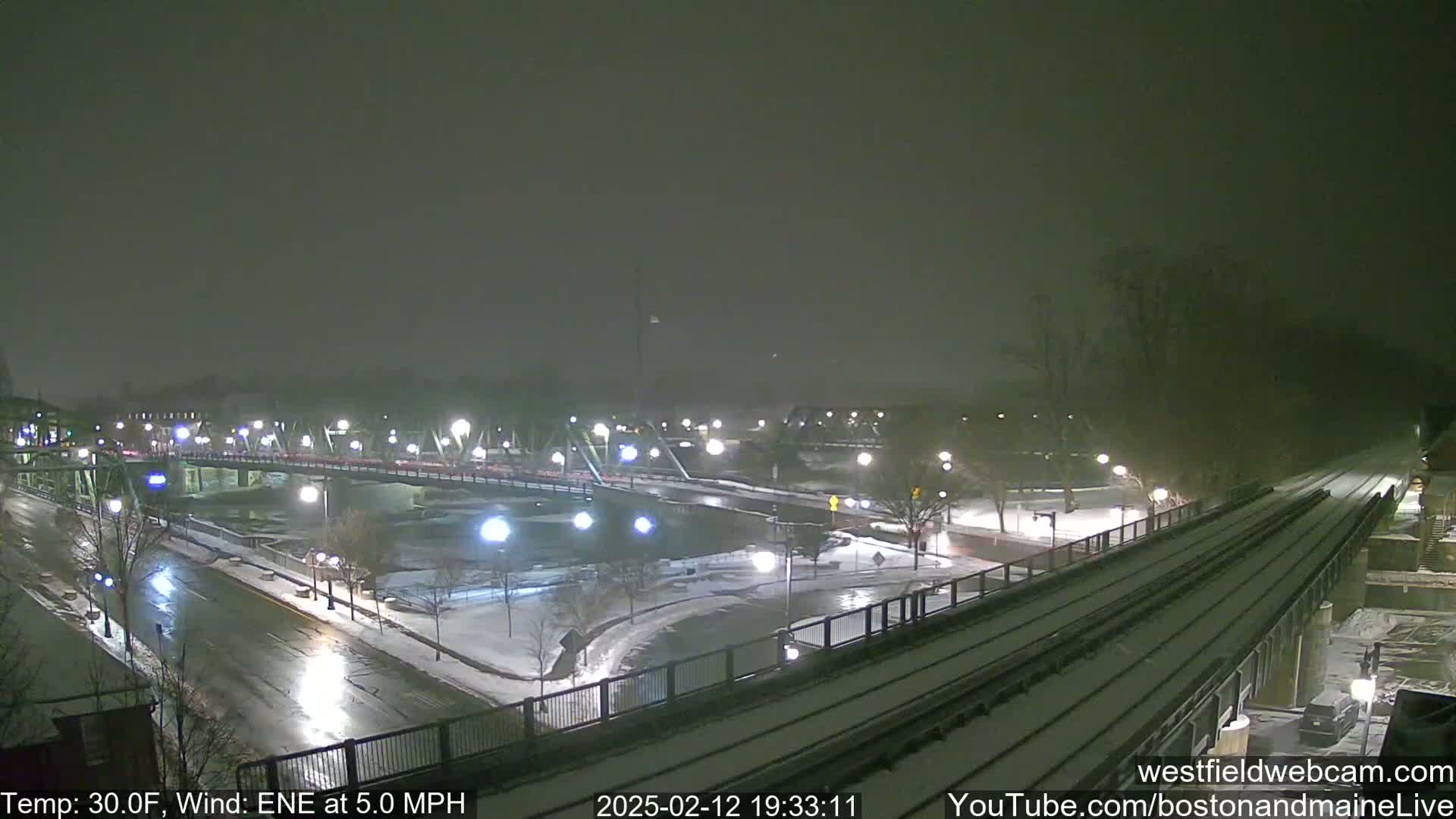 A snow-covered nighttime scene shows a bridge over a river with cars driving on it and a train track alongside a road.