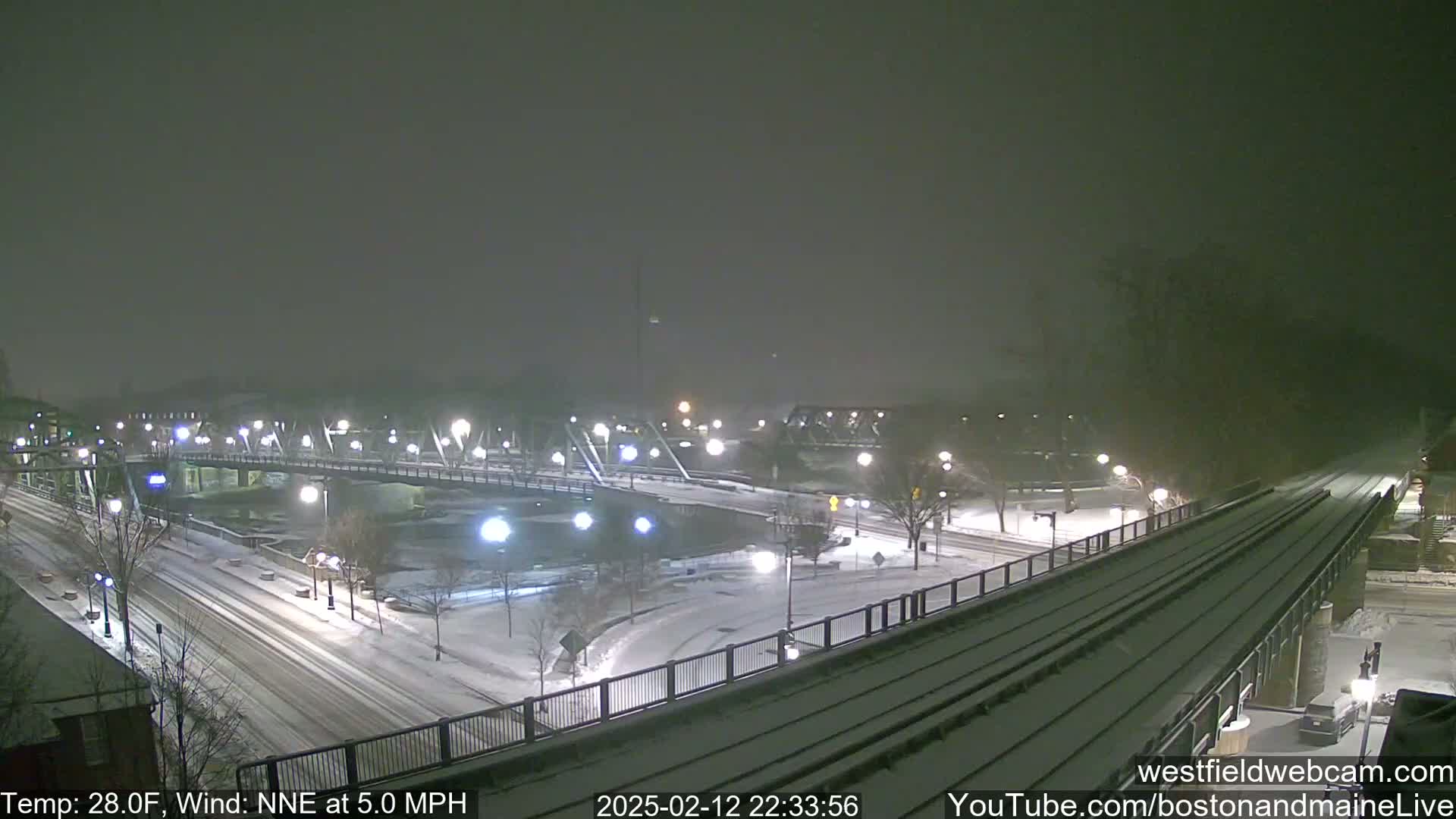A snow-covered bridge and roadway are seen at night, under light snowfall with a 28°F temperature and a 5.0 MPH wind from the north-northeast.