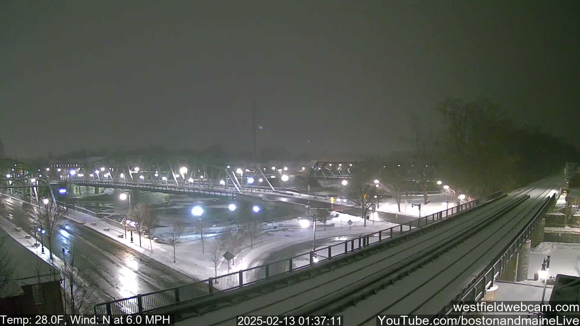 A snow-covered bridge and roadway are visible at night, under a falling snow.