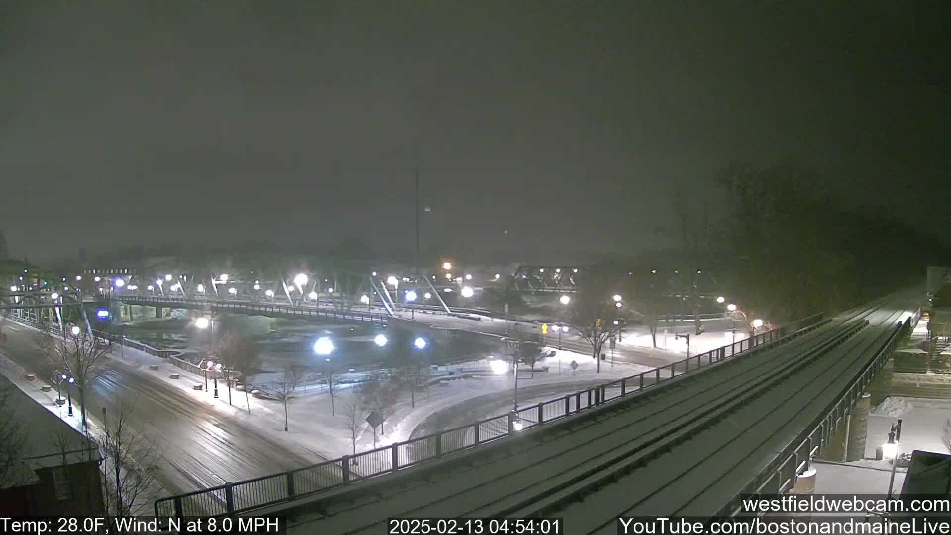 A snow-covered bridge and roadway are visible under a dark, snowy night sky, with a train track running alongside.