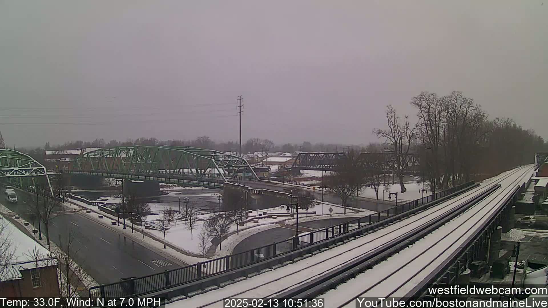 A snow-covered train track and two bridges span a river in a lightly snowing, overcast scene.