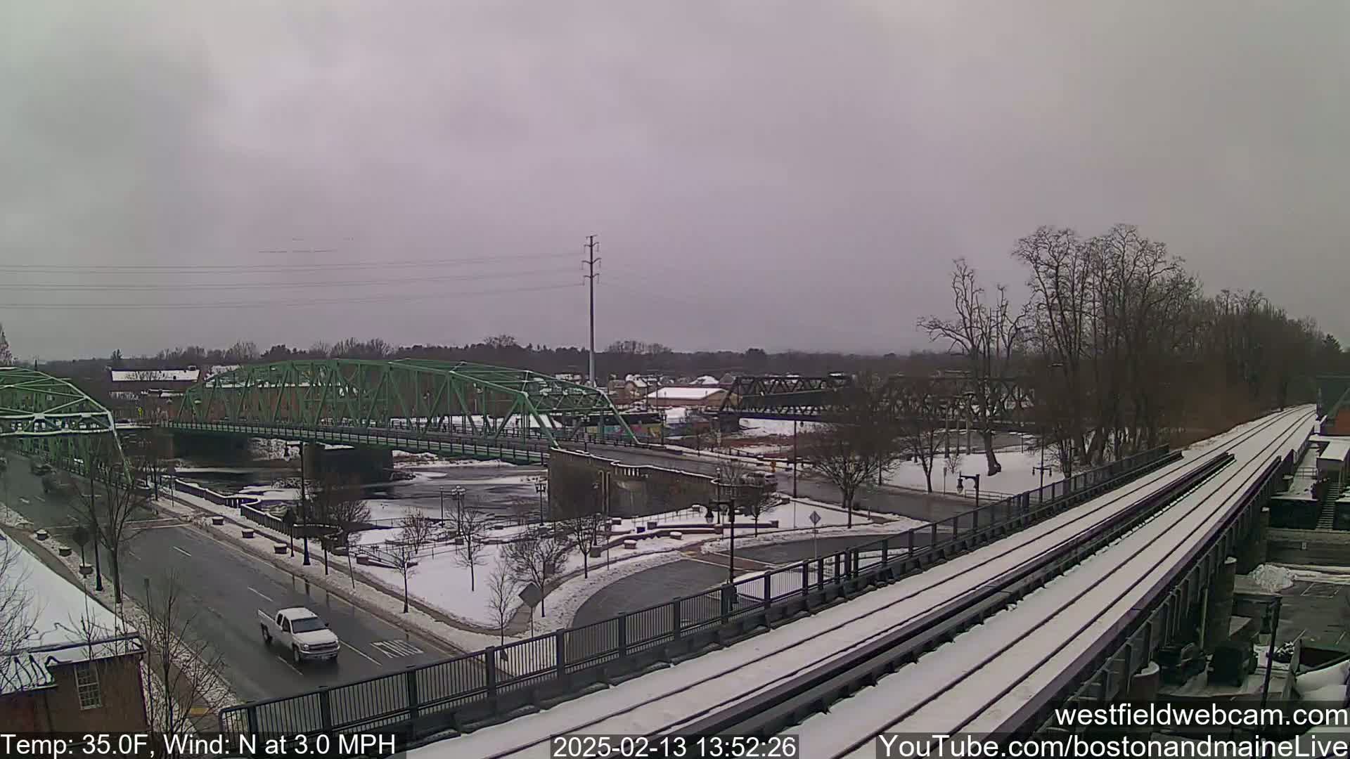 A snow-covered landscape shows a green metal bridge over a river, a roadway with a white truck, and train tracks leading into the distance under an overcast sky.