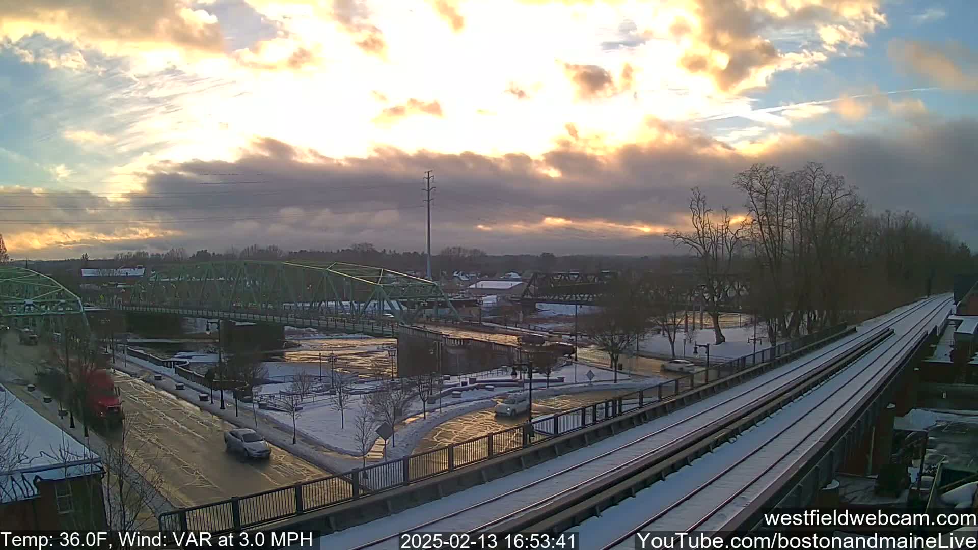 A partly cloudy sunset illuminates a snow-dusted town scene featuring a river, two bridges, and train tracks.