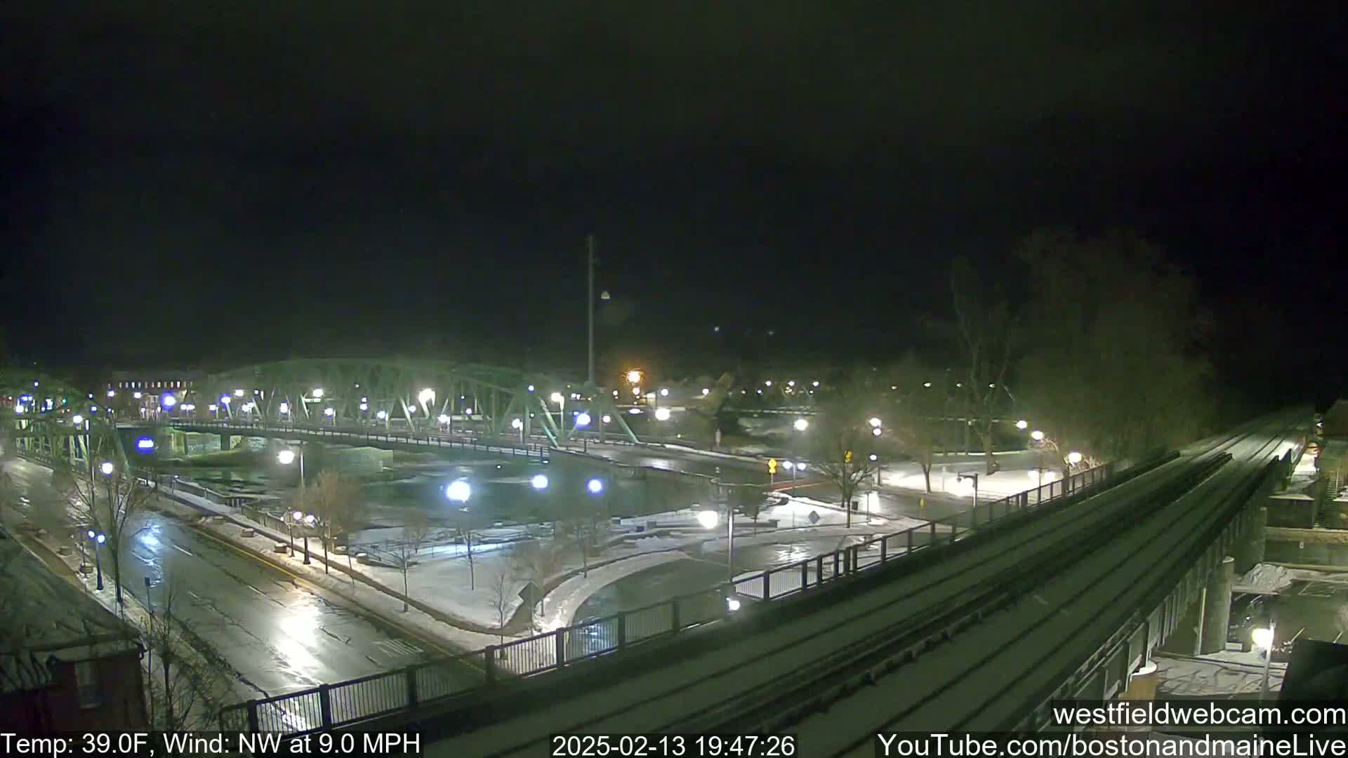 A nighttime view of a snow-covered bridge, roads, and train tracks alongside a river, under a dark sky with a northwest wind.