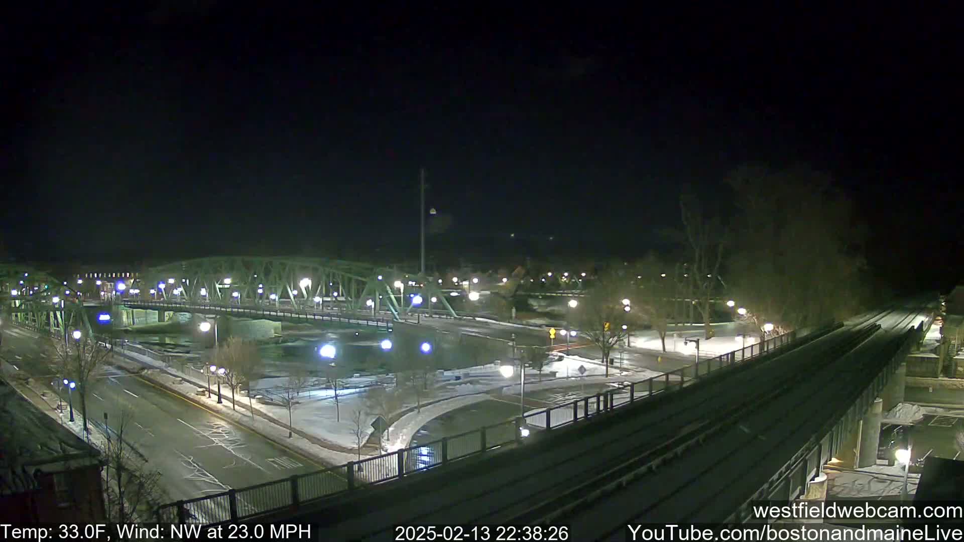 A nighttime view of a snow-covered bridge and roadway intersecting with a train track, under a dark sky with a northwest wind at 23 mph and a temperature of 33 degrees Fahrenheit.