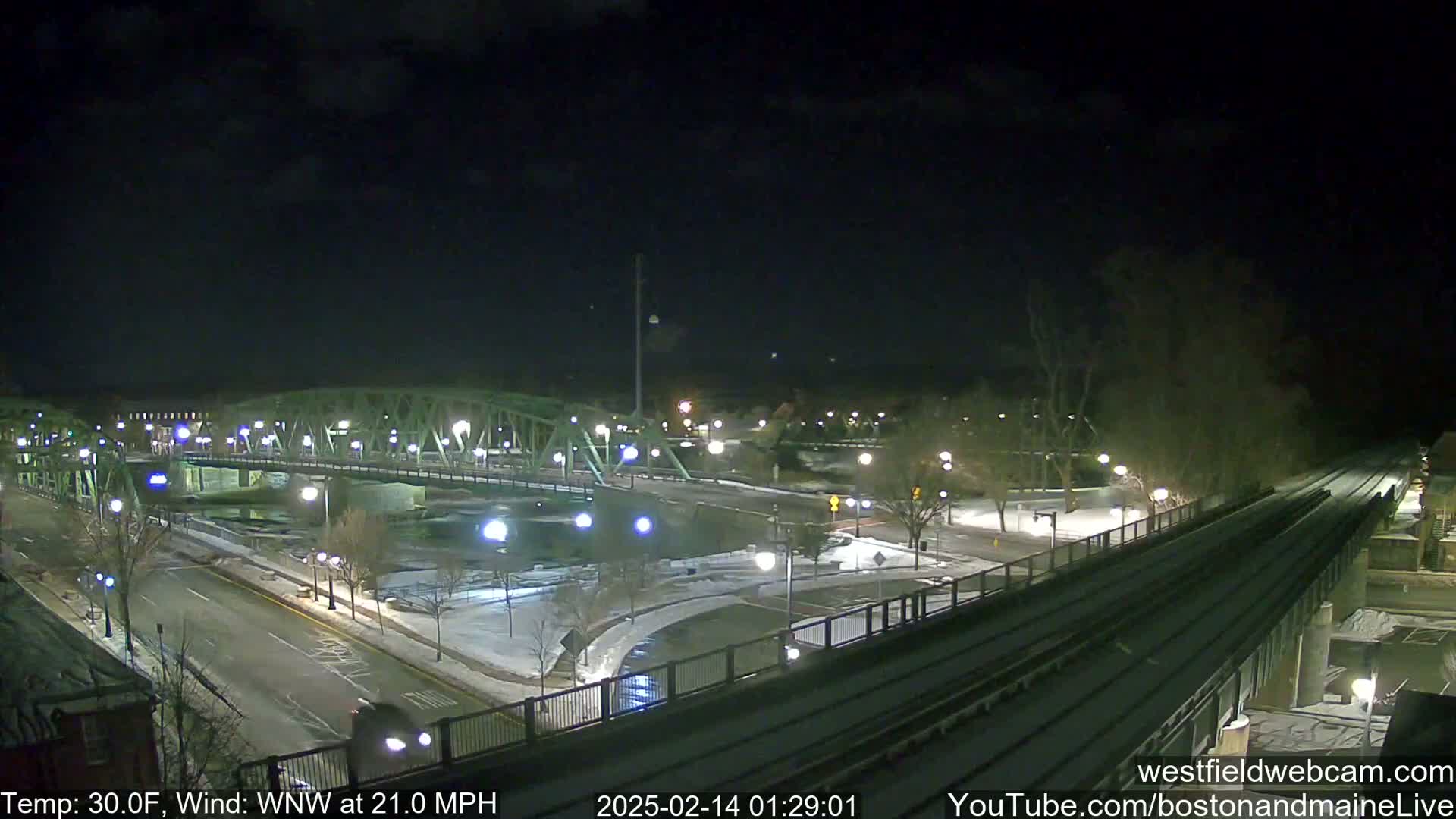 A nighttime, snow-covered scene shows a bridge over a river, roads, and train tracks under a dark sky with a wind of 21 mph.
