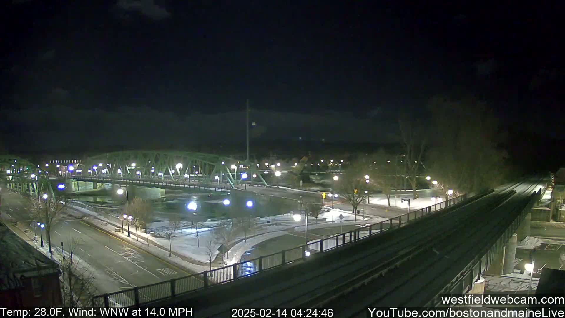 A nighttime view of a snow-covered bridge and roadway, with a train track running alongside, under a mostly clear sky.