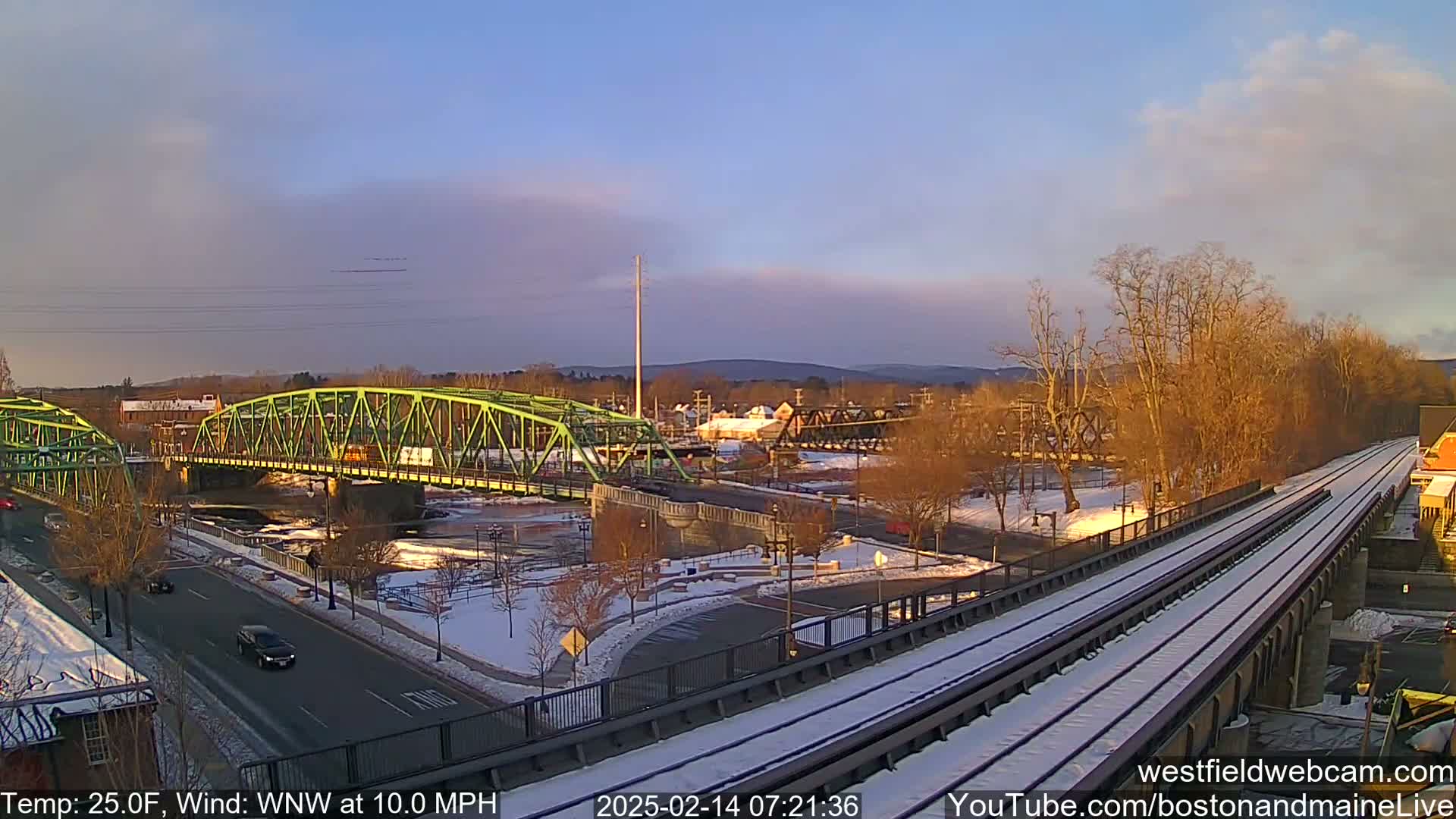 A snow-covered train track runs alongside a river, crossing two bridges under a partly cloudy, sunrise sky.