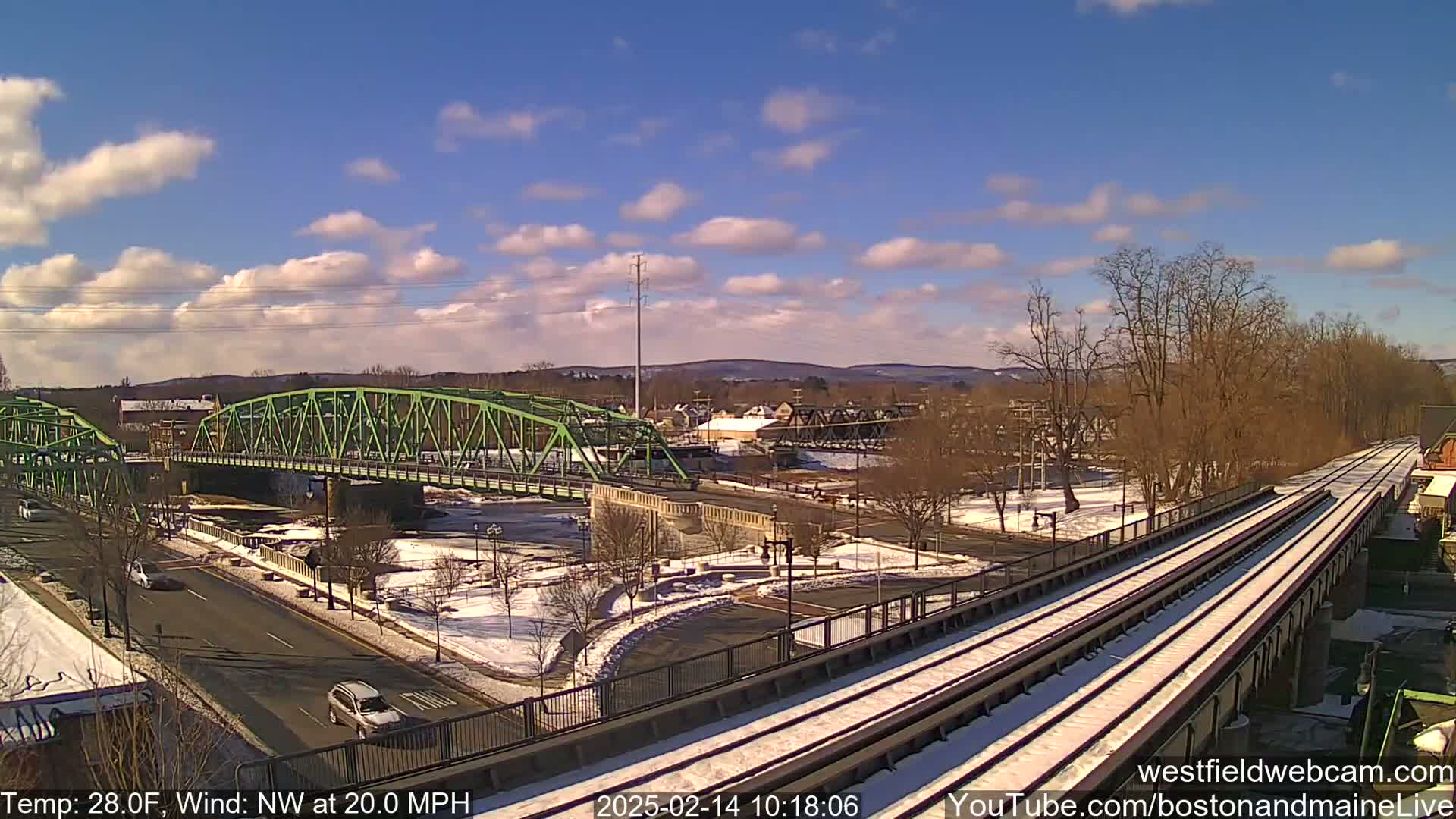 A green bridge spans a river in a partly snowy town under a mostly sunny sky with a northwest wind blowing at 20 mph.