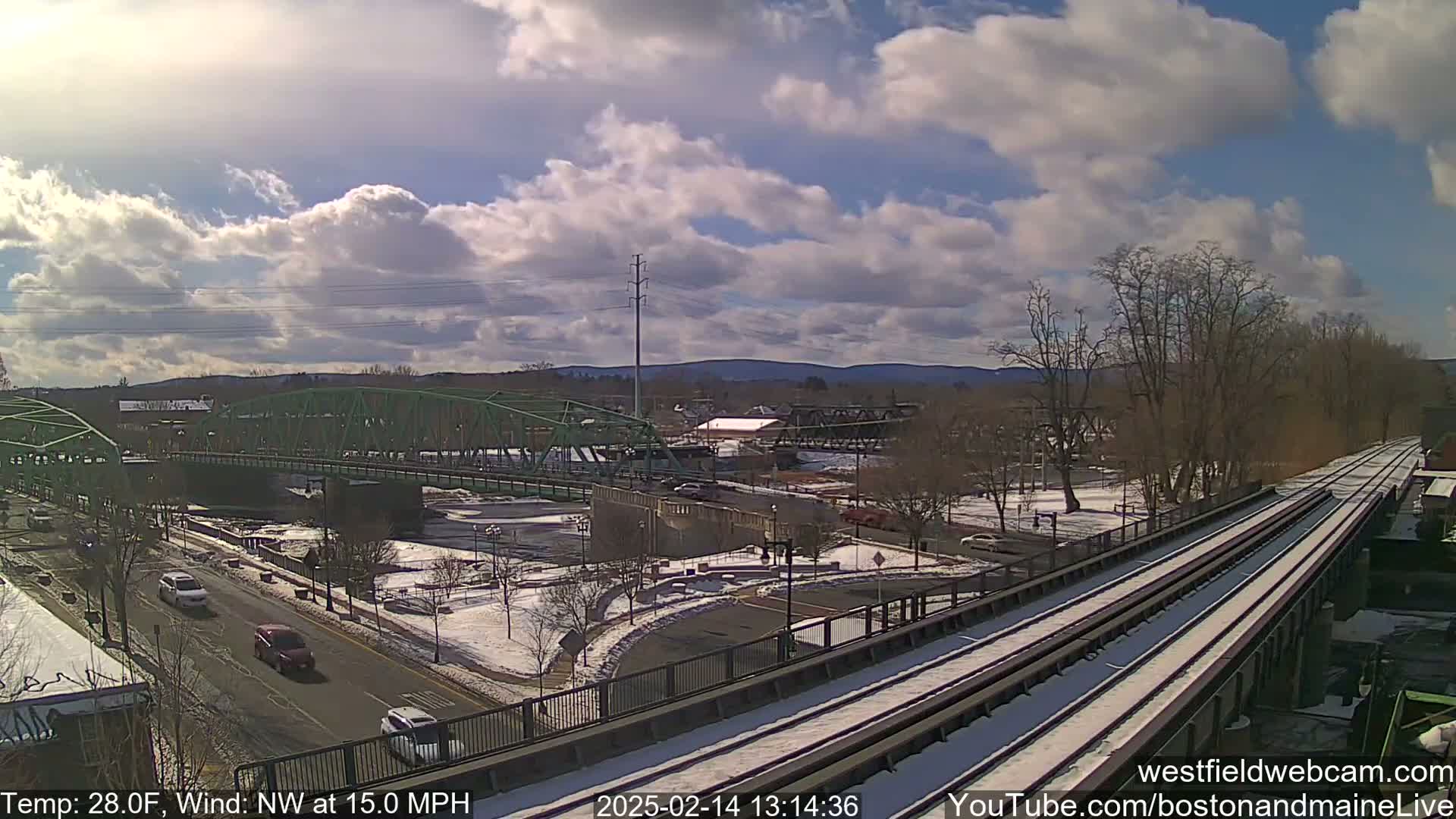 A partly cloudy, cold day with light snow on the ground and train tracks, overlooking a river and green bridge with cars driving on a road below.