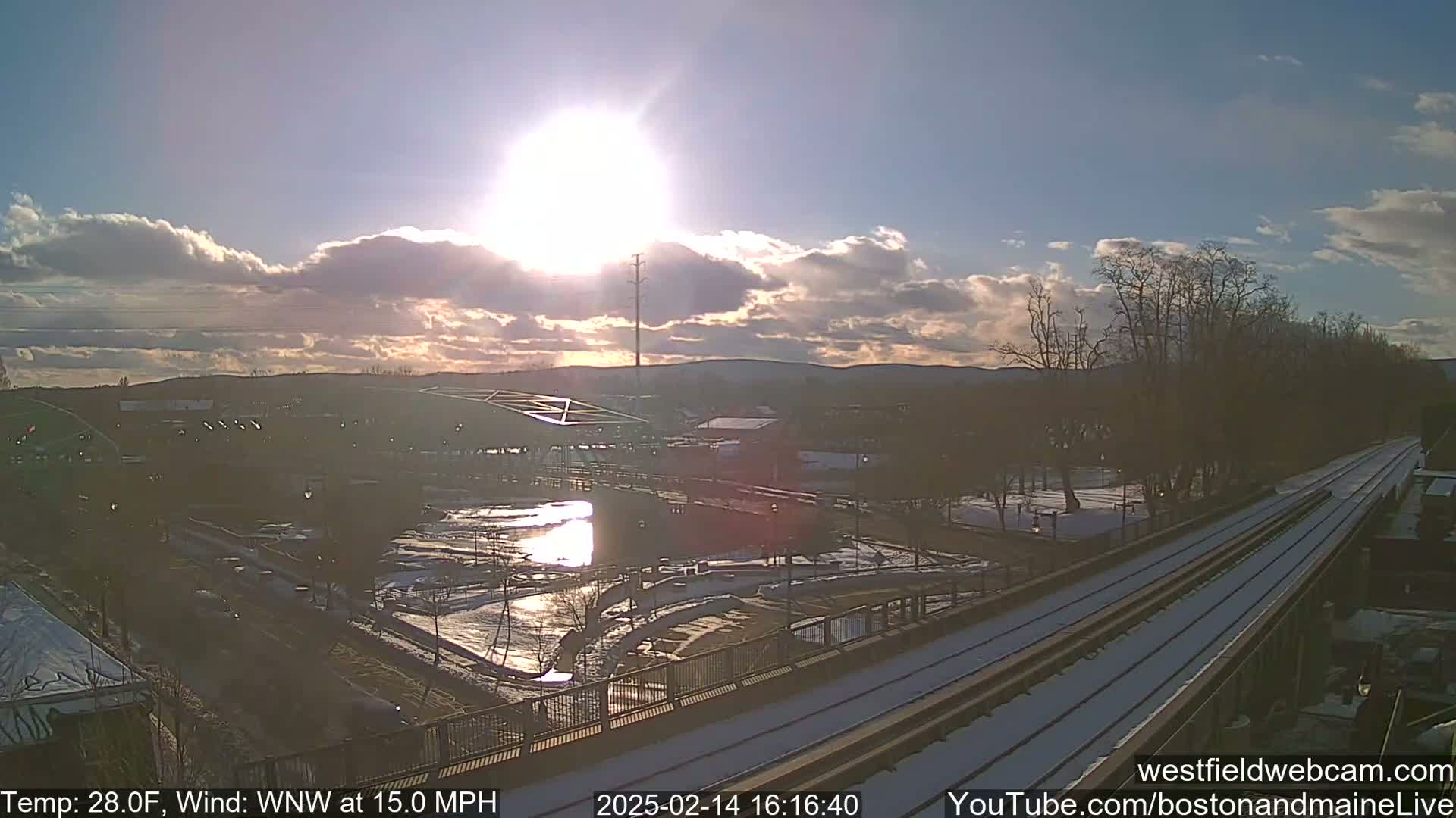 A partly cloudy sunset illuminates a snow-dusted park and train tracks near a river, with a bridge in the background and a wind of 15 mph.