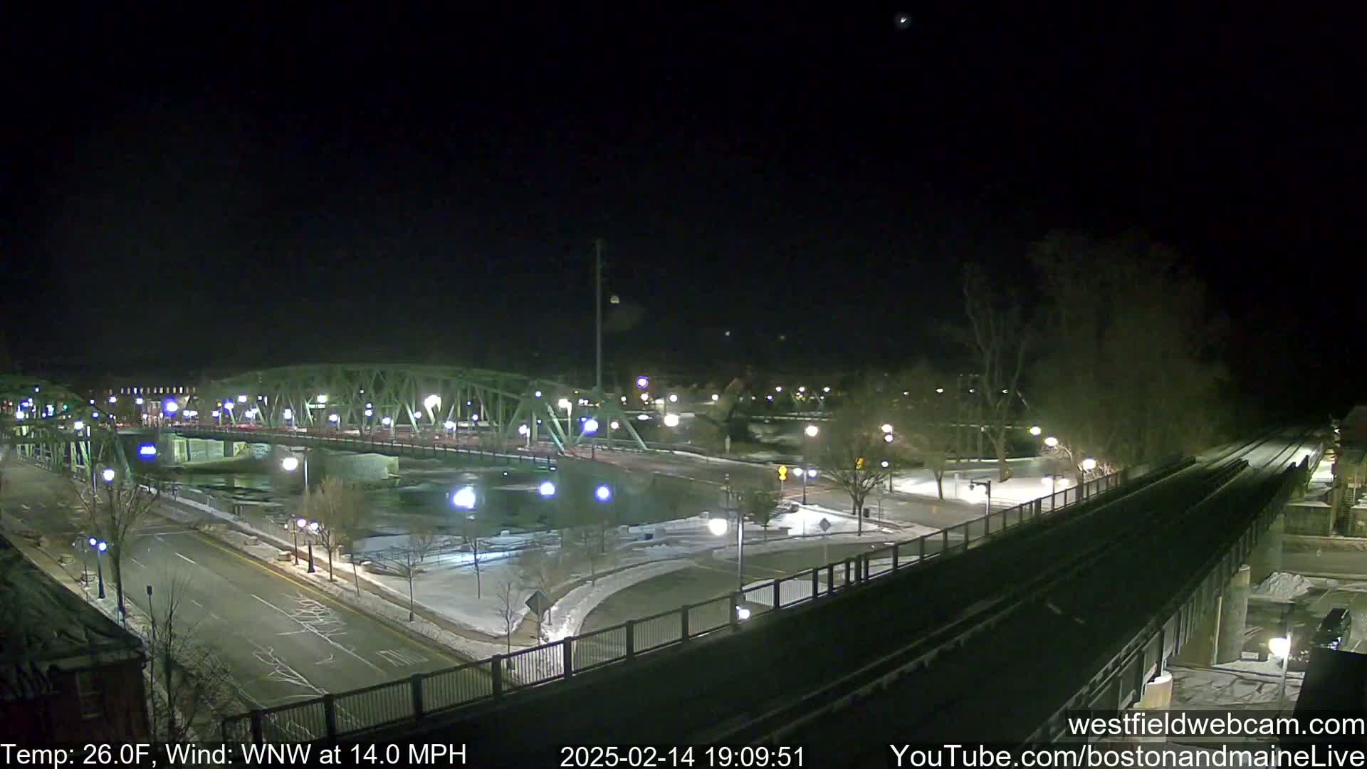 A nighttime view of a bridge over a river, with snow on the ground and a 26°F temperature and a 14 mph wind from the west-northwest.