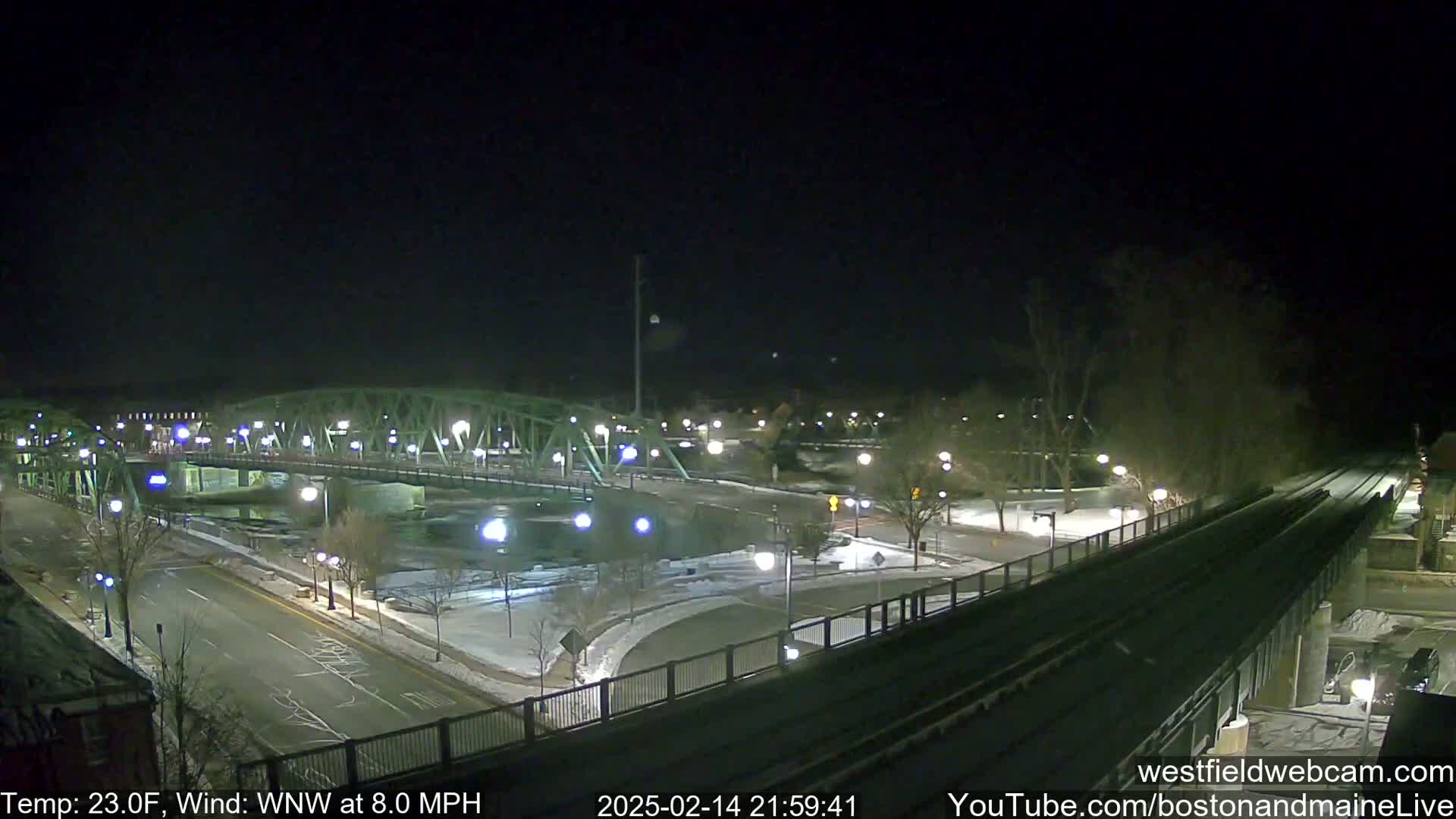 A nighttime view of a bridge over a river, with snow on the ground and a temperature of 23 degrees Fahrenheit and a wind of 8 mph from the west-northwest.