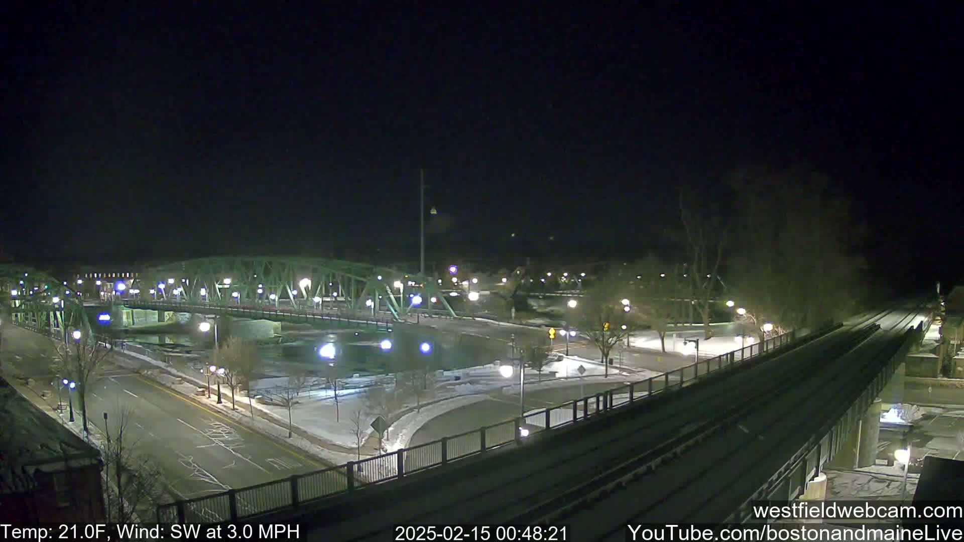 A snow-covered bridge and roadway are illuminated at night under a dark sky, with a light southwest wind.