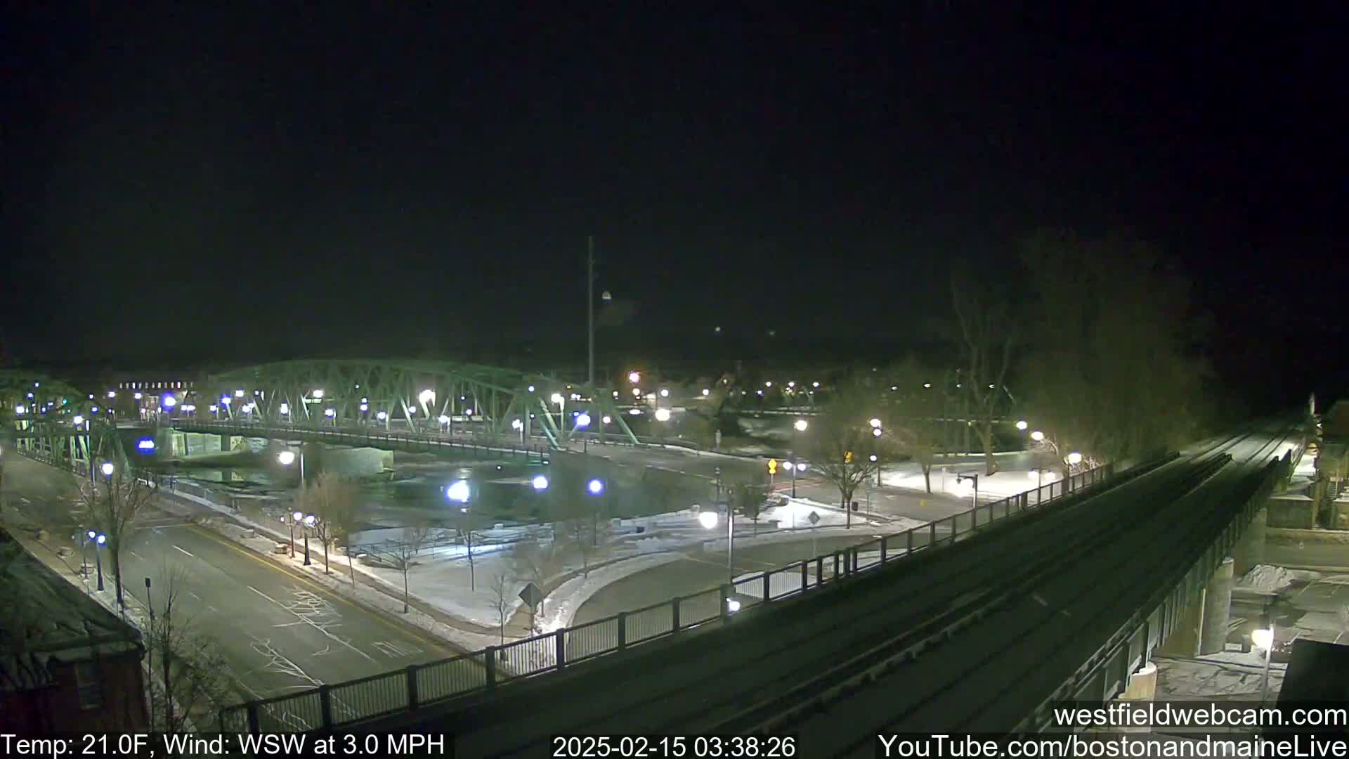 A nighttime view of a snow-covered bridge and roadway, illuminated by streetlights, with a light wind.