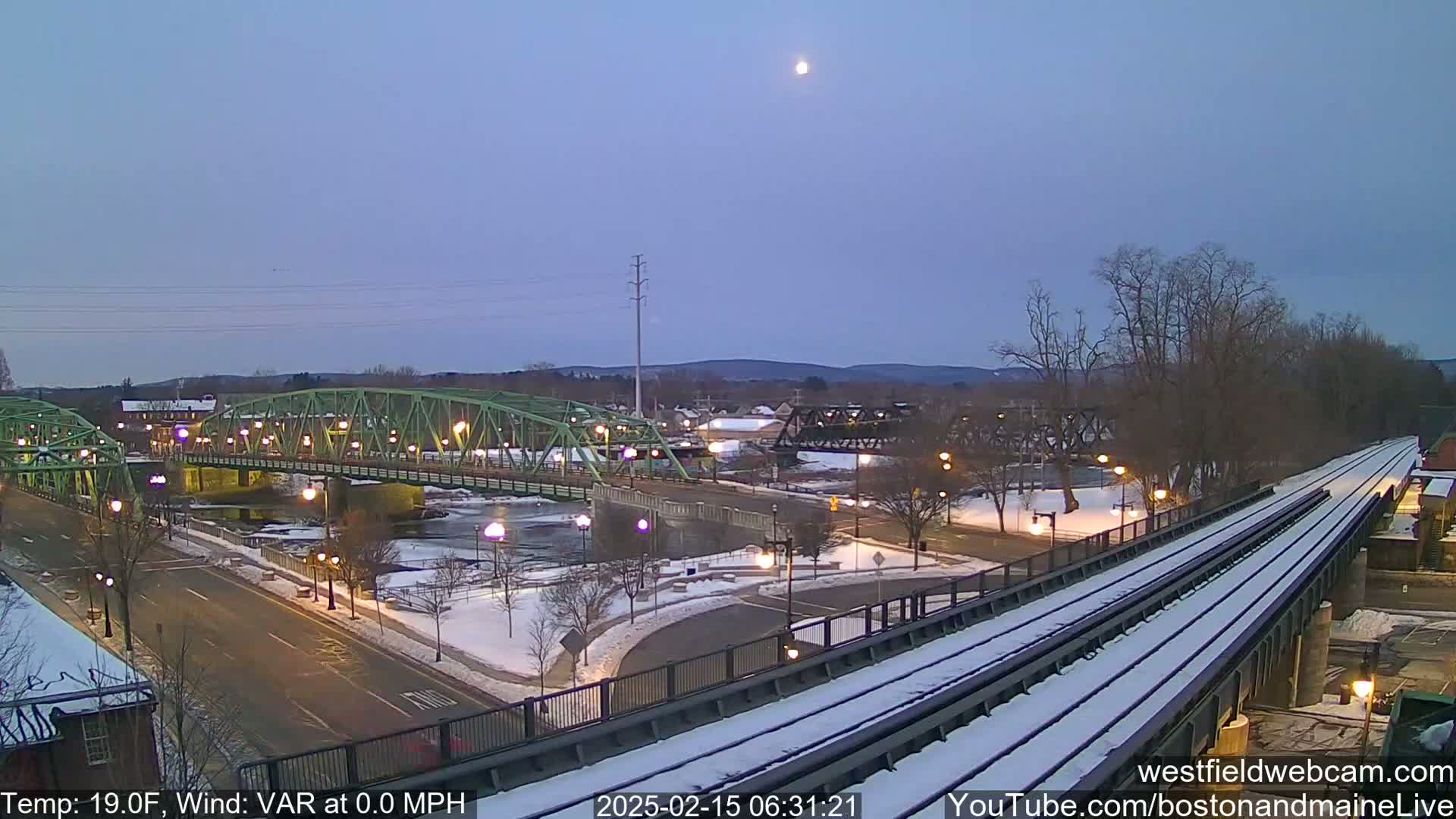A snow-covered train track overlooks a green bridge spanning a river, with a town in the background under a twilight sky with a visible moon.