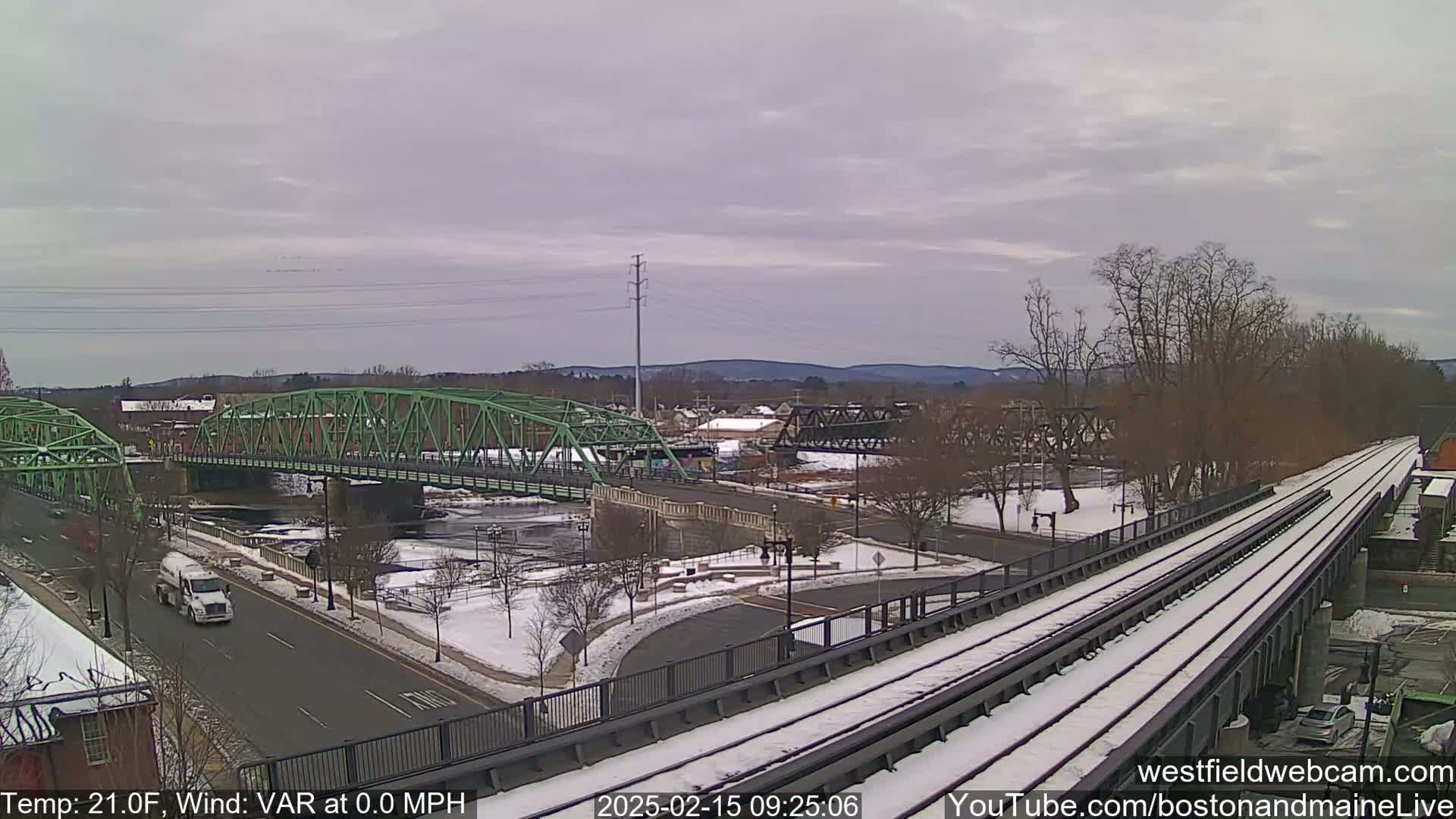 A snow-covered train track overpasses a river with two bridges and a road, under a cloudy sky.