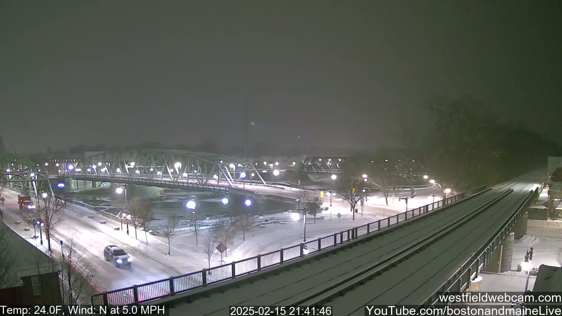 A snow-covered bridge and roadway are seen at night, under a light snowfall, with a few vehicles traveling across.