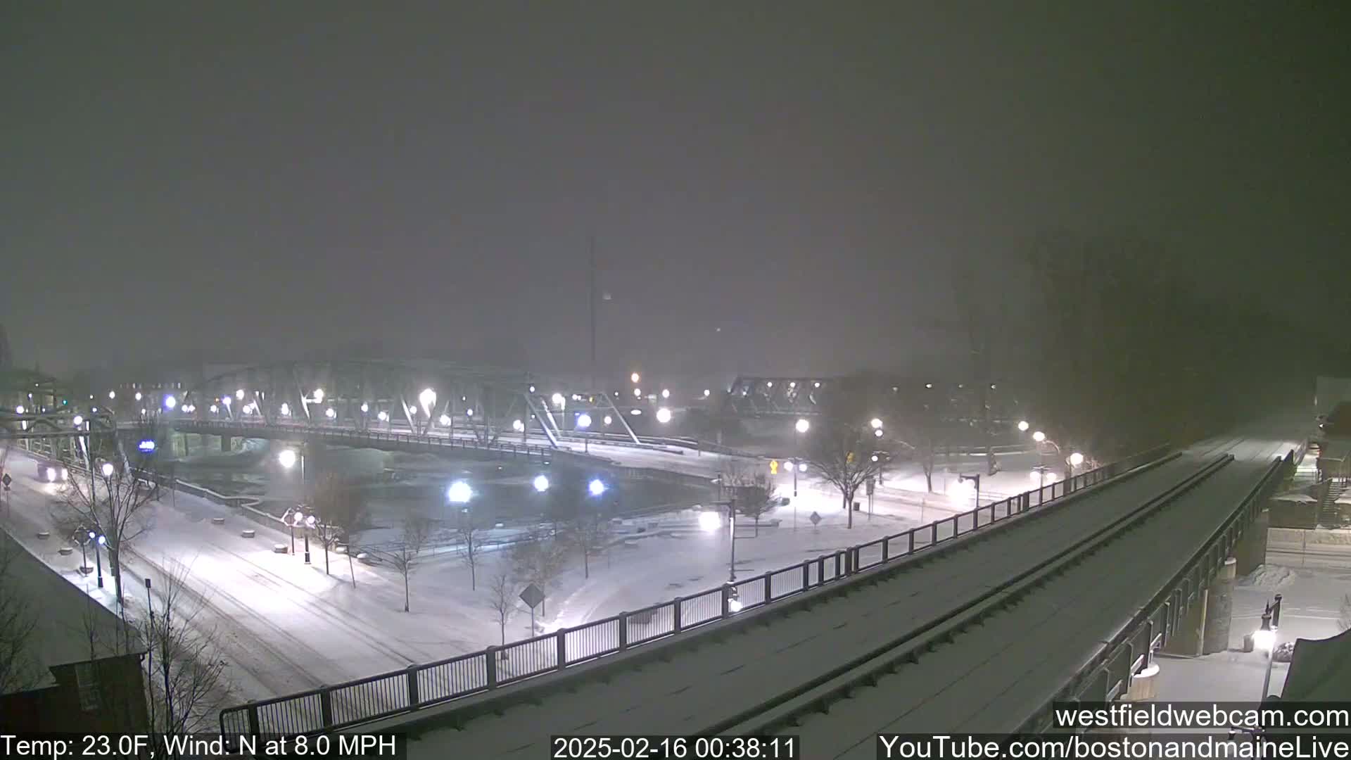 A snow-covered bridge and roadway are visible under a nighttime snowfall, with a train track running alongside.