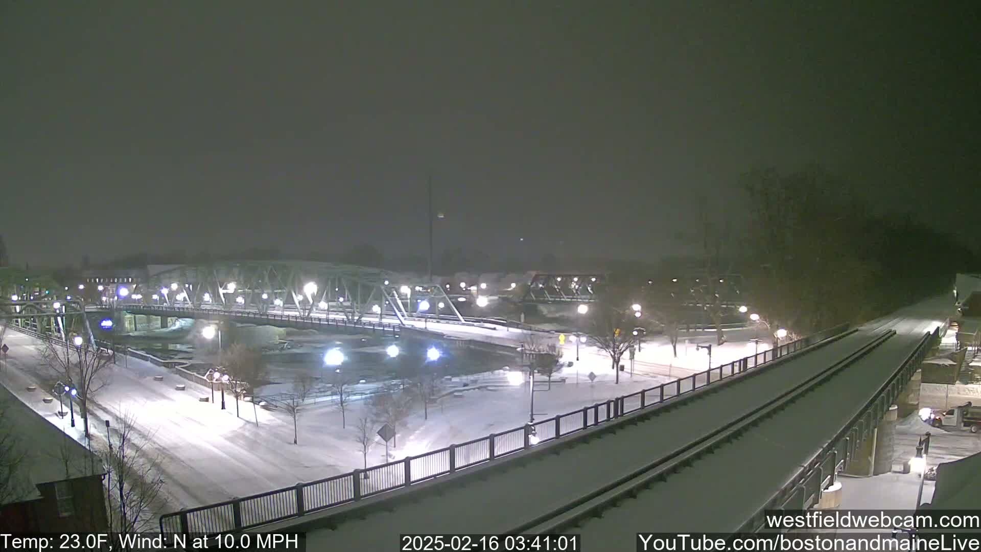 A snow-covered bridge and roadway are illuminated at night by streetlights, with a train track alongside, under a lightly snowing sky.