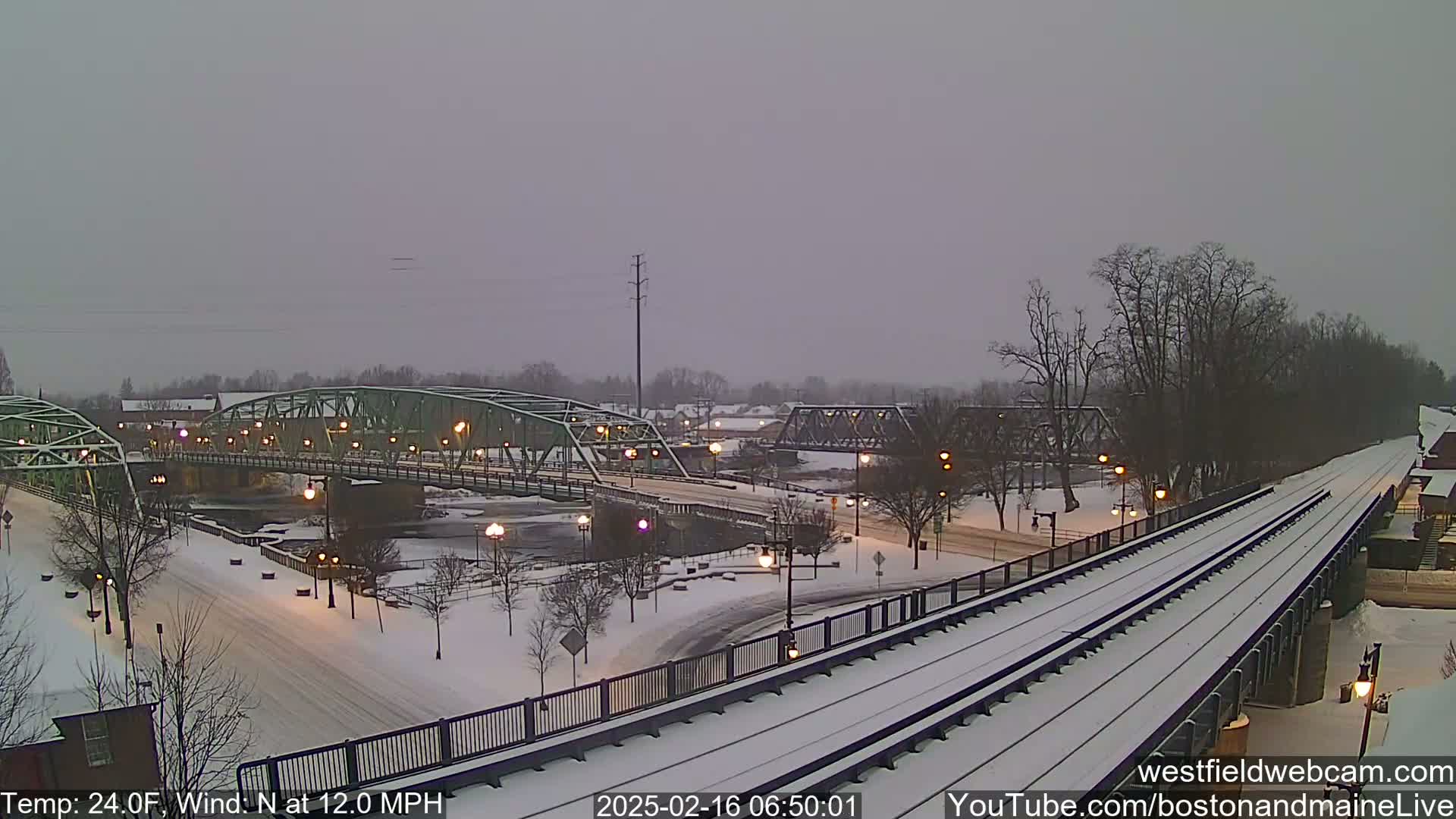 A snow-covered town scene with two bridges over a river, train tracks, and streetlights under a cloudy sky.