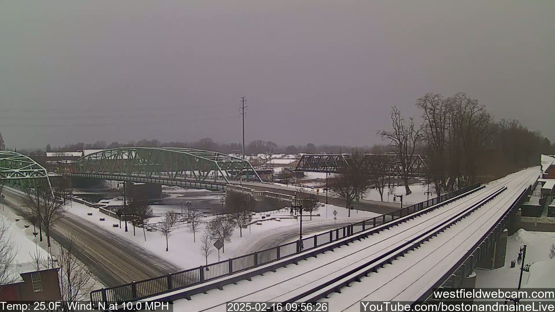 A snow-covered town scene shows two bridges over a river, with train tracks in the foreground and a light snowfall.