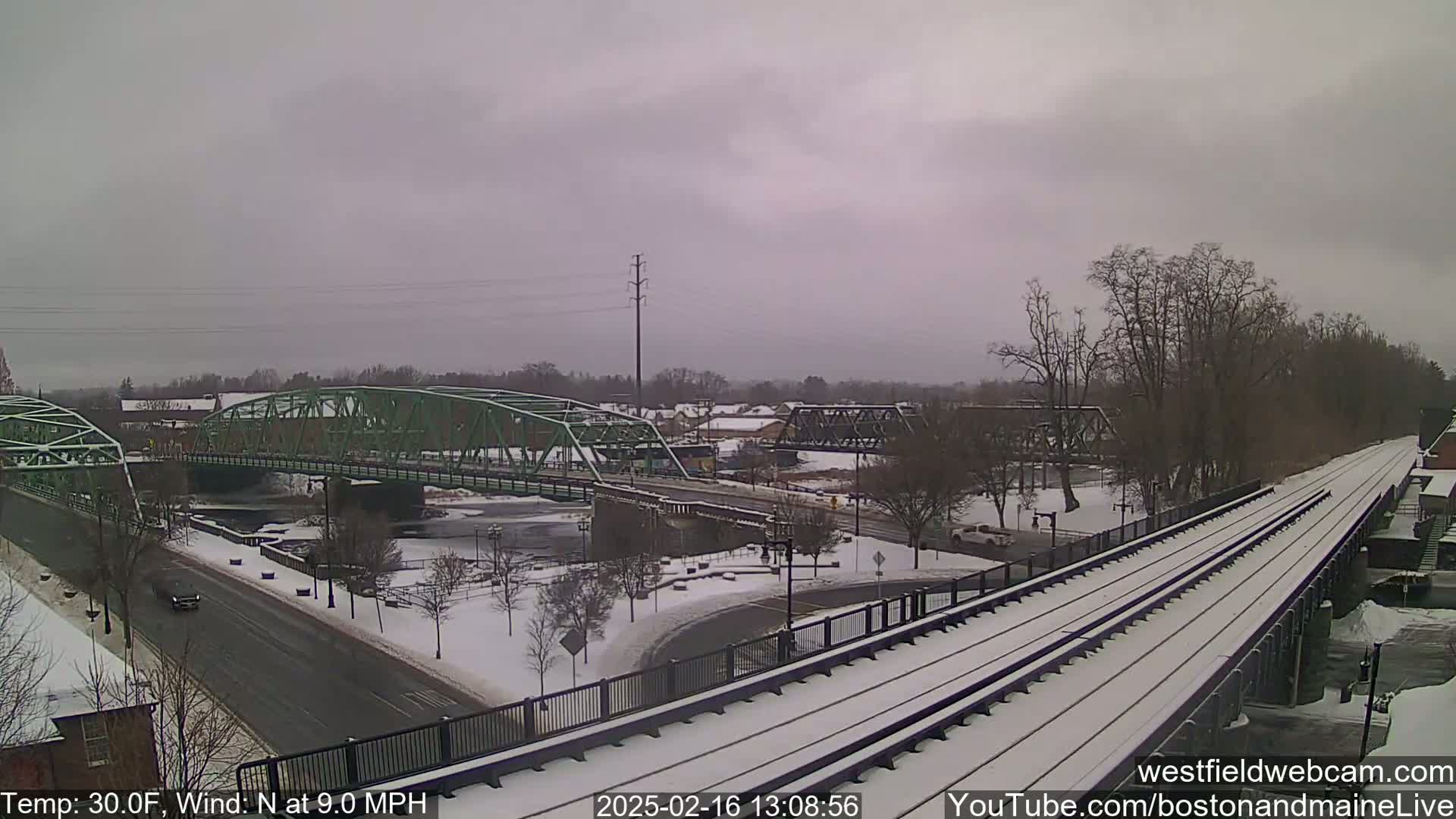 A snow-covered town scene shows two bridges over a river, with train tracks and a road in the foreground under a gray sky.