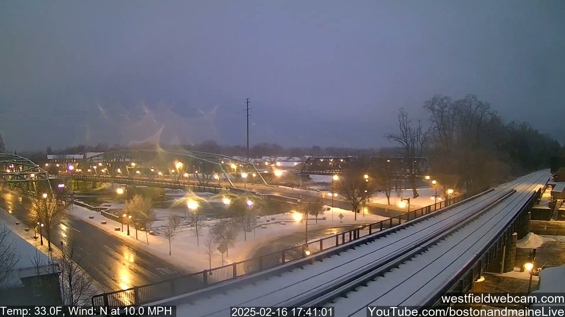 A snow-covered town scene at twilight, showing a bridge, roads, train tracks, and a river, with a north wind blowing at 10 mph and the temperature at 33°F.