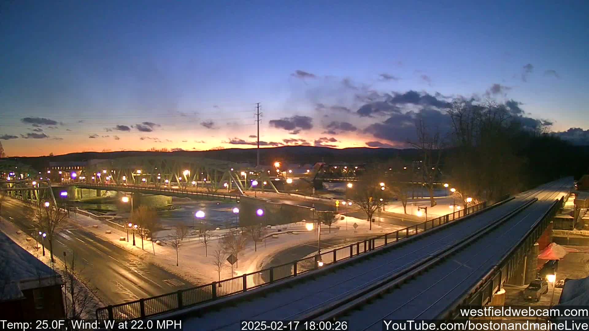 A snow-covered city scene at twilight shows a bridge over a river, roadways with streetlights, and a train track in the foreground, with a wind of 22 mph and a temperature of 25 degrees Fahrenheit.