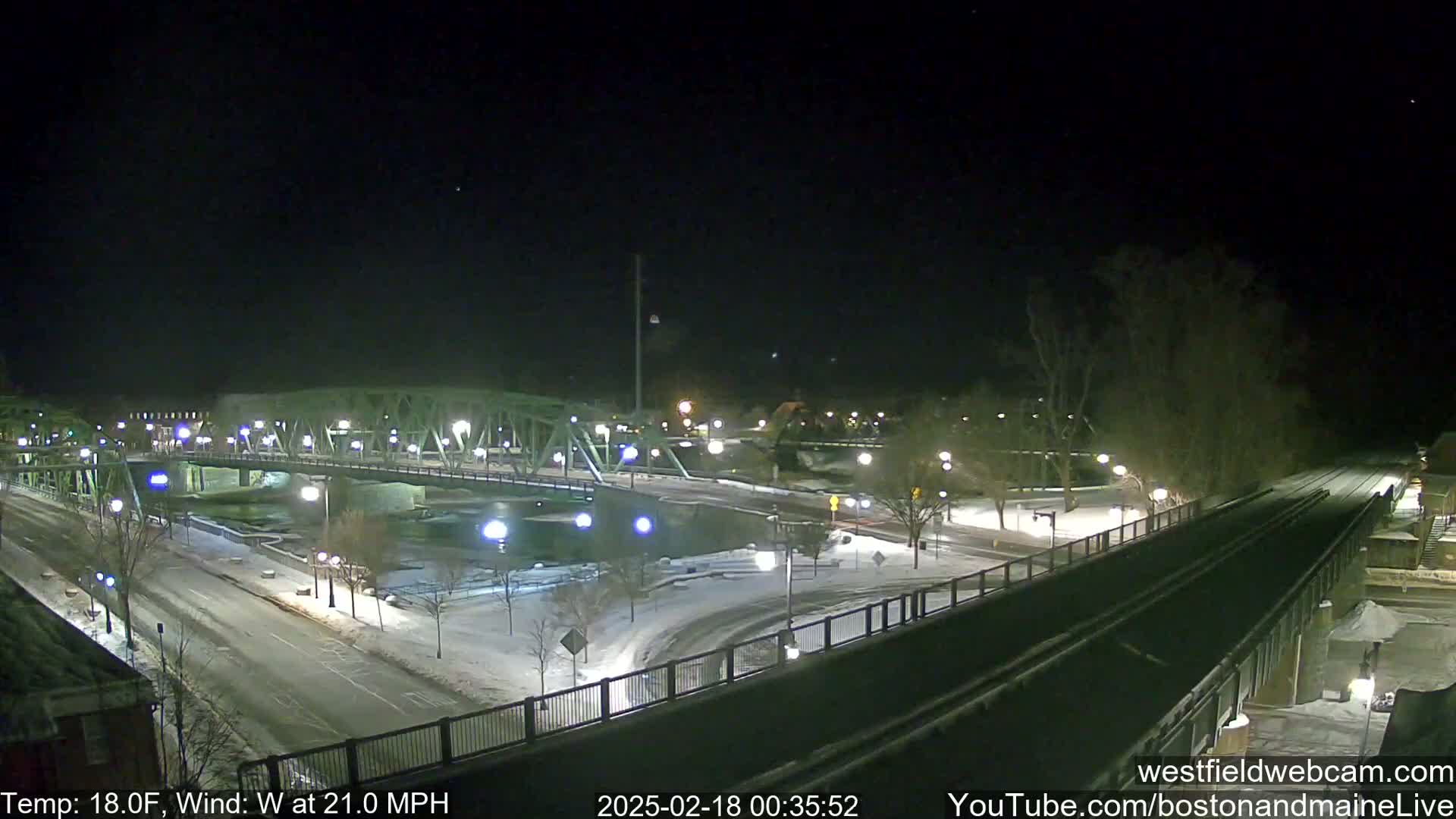 A snow-covered bridge and roadway are illuminated at night, with a river visible below, under windy, 18-degree Fahrenheit conditions.