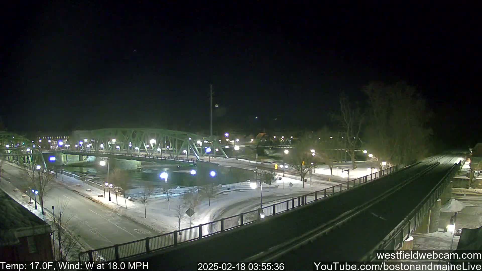 A snow-covered nighttime scene shows a bridge over a river, roads, and surrounding buildings under a dark sky with a 17°F temperature and 18 mph wind.