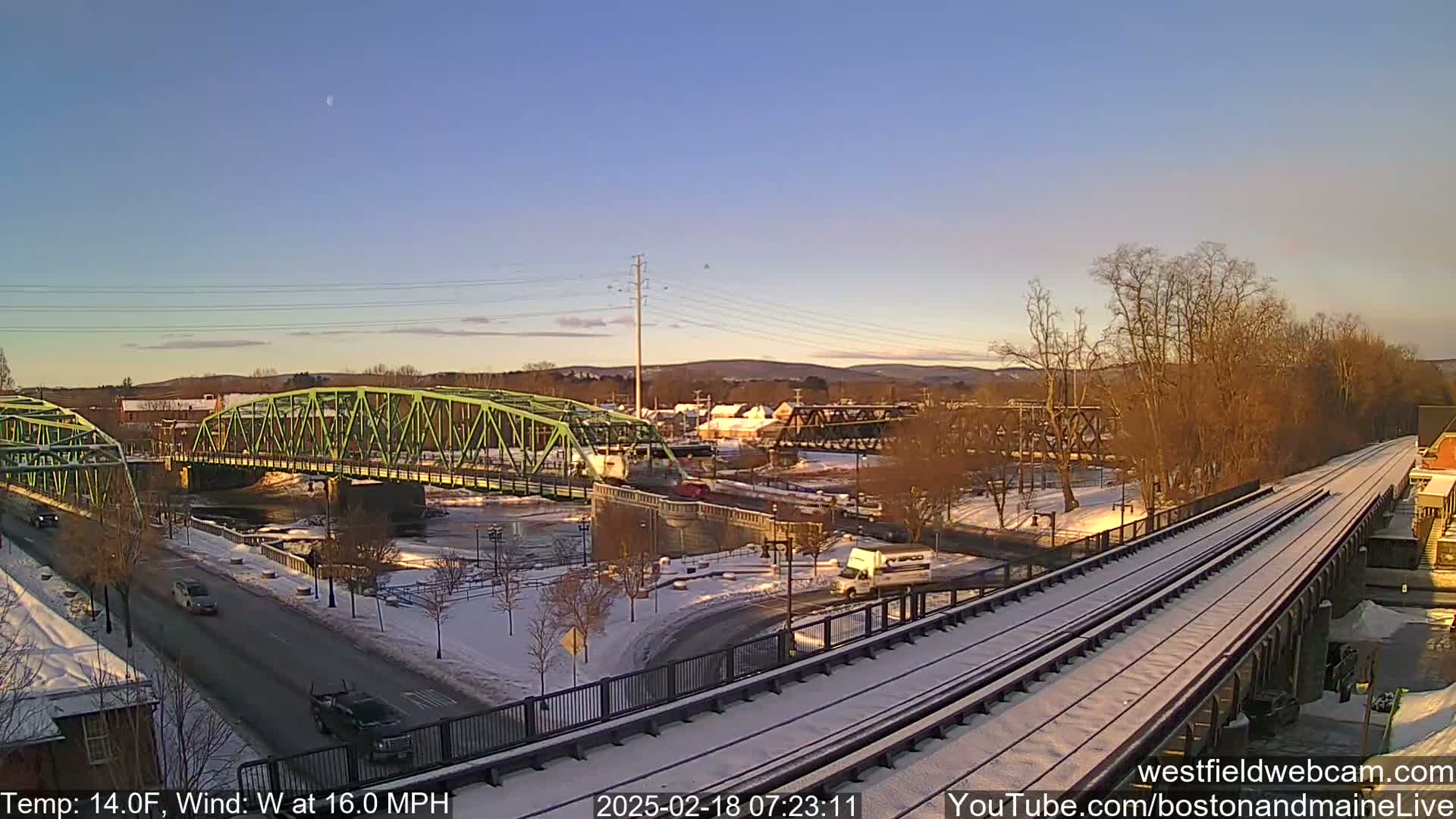 A snow-covered landscape with a light green bridge over a river, a train track, and a partially visible town under a clear, light-blue sky.