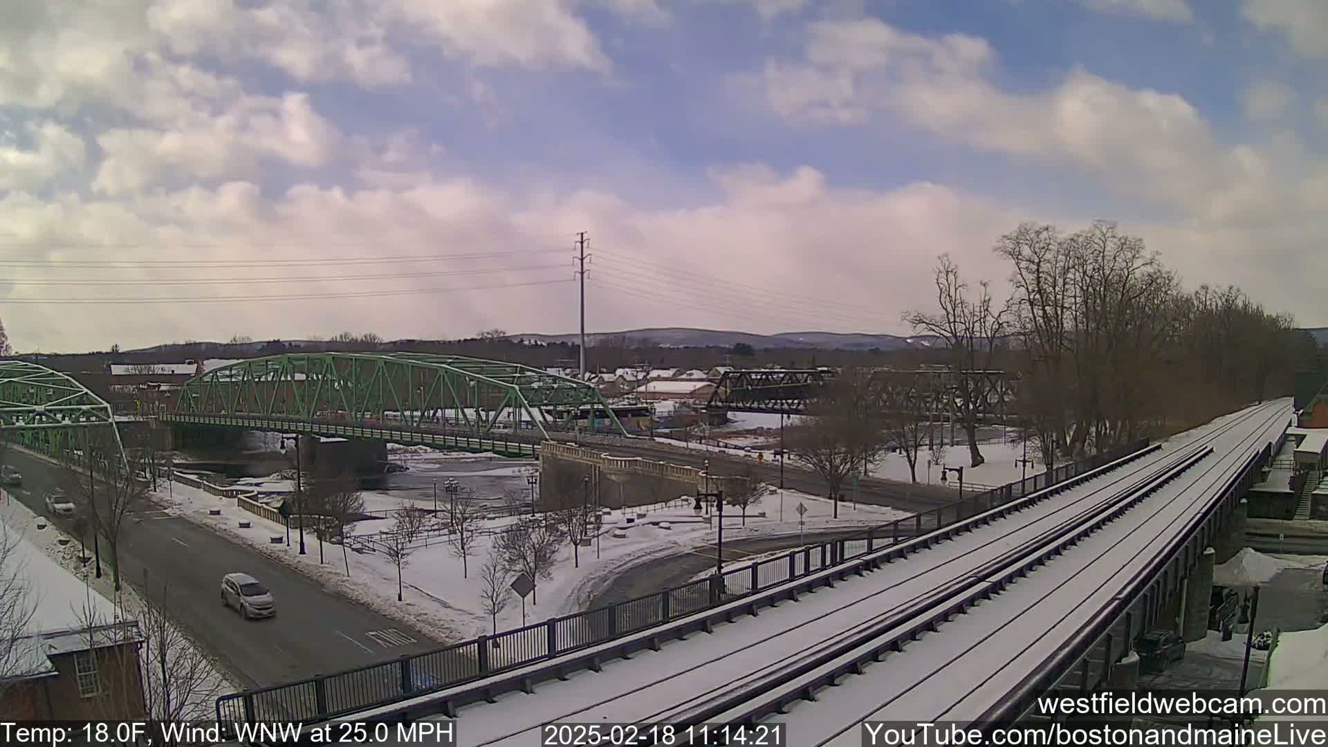 A snow-covered train track stretches across the foreground, with a green bridge and a river visible in the background under a partly cloudy sky.