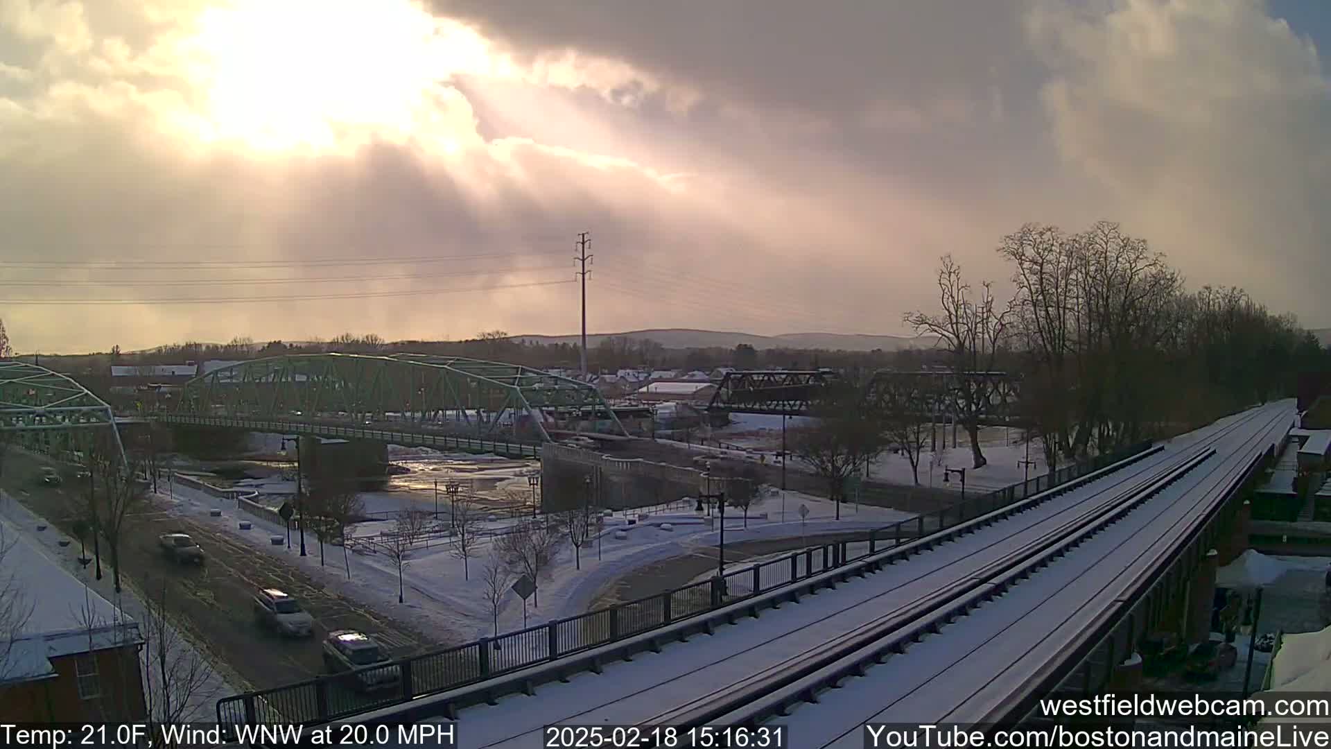 A snow-covered landscape with two bridges over a river, train tracks, and cars driving on a road under a partly cloudy sky with sun rays breaking through the clouds.