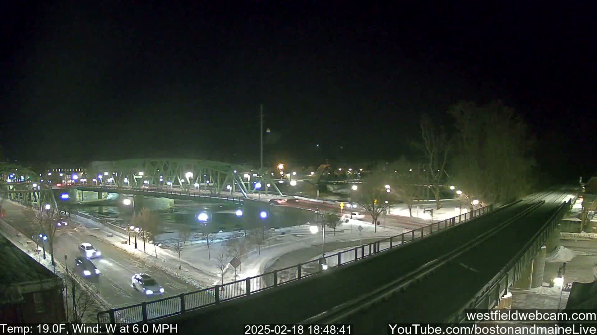 A nighttime view of a snow-covered bridge and roadway with several cars driving, under a clear, dark sky with a temperature of 19°F and a 6.0 mph wind.