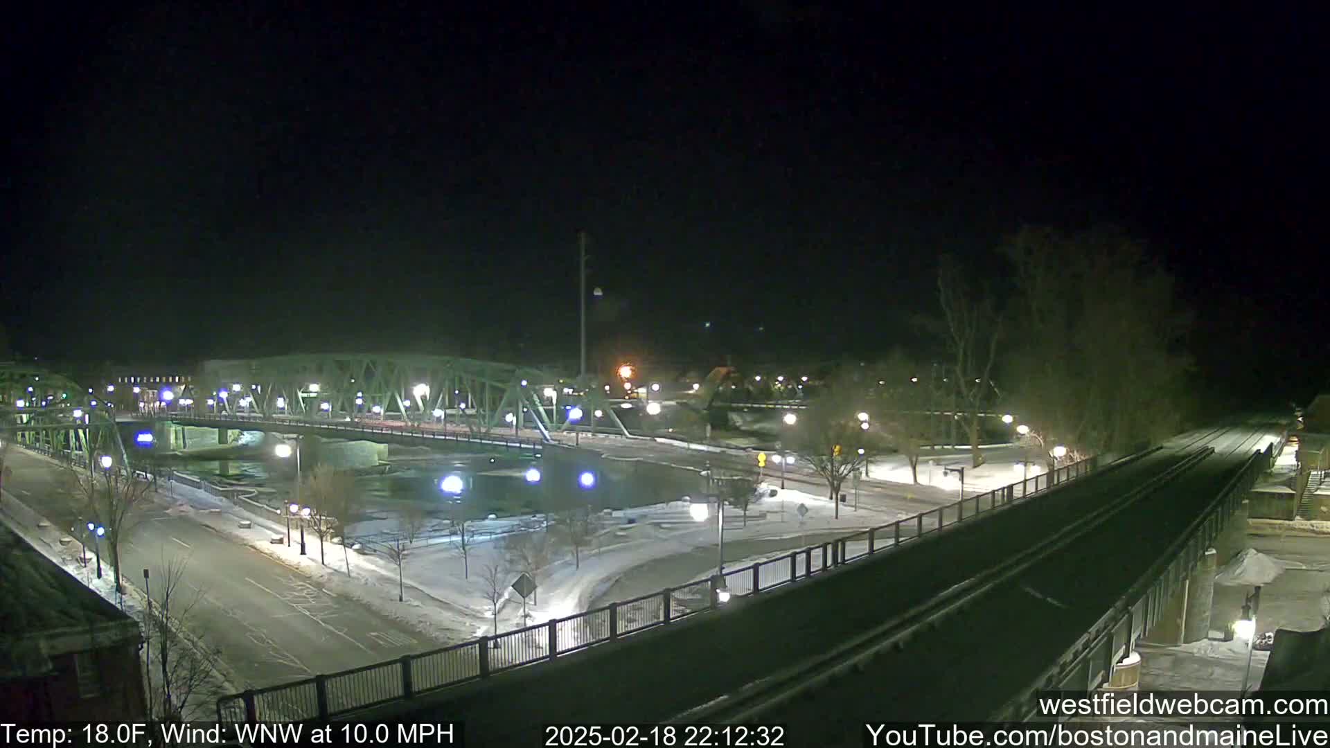 A nighttime view of a snow-covered town with a bridge over a river, under a clear, dark sky.