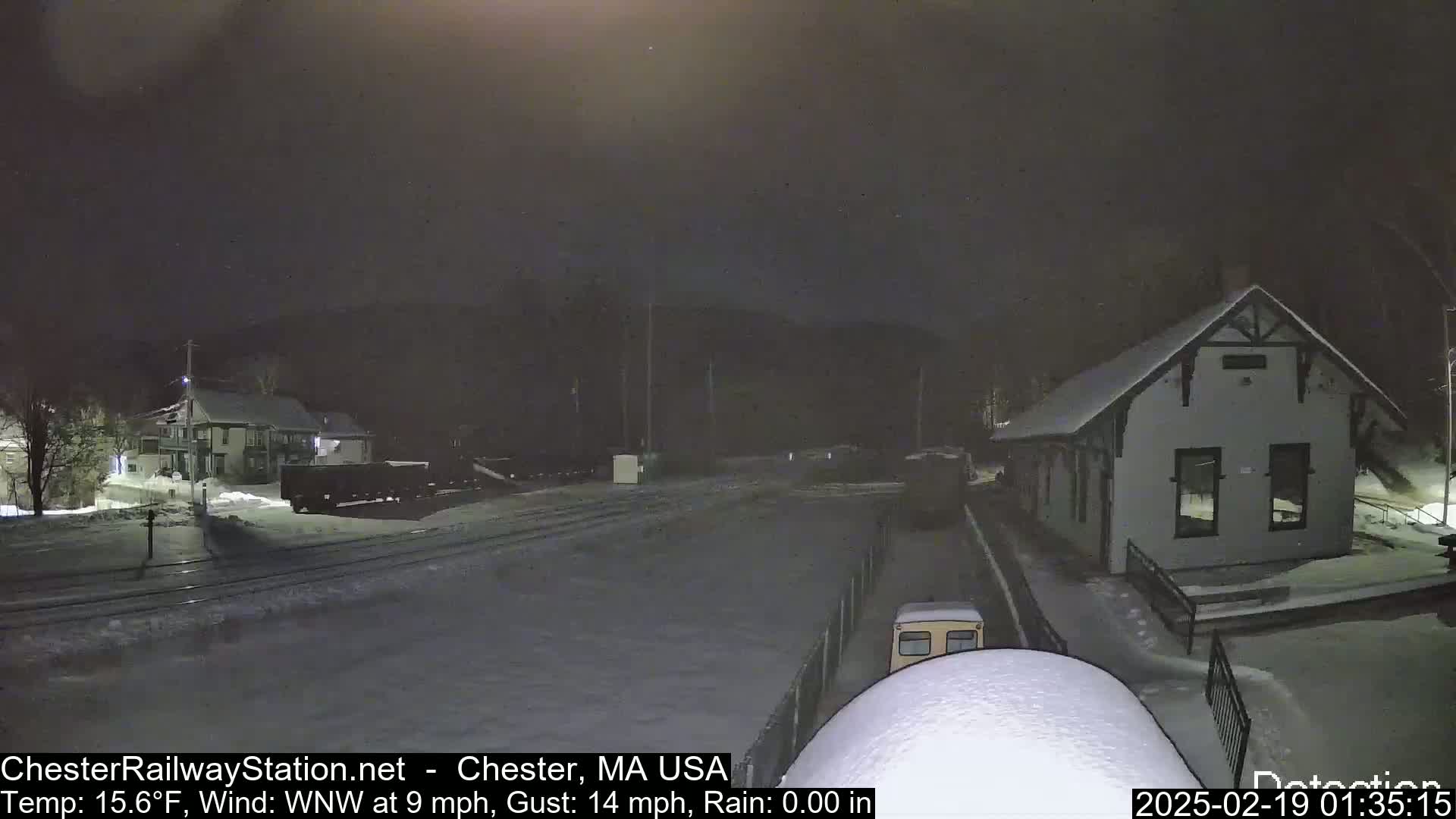 A snow-covered train station and surrounding area are visible at night under a dark sky, with light rain or snow falling.