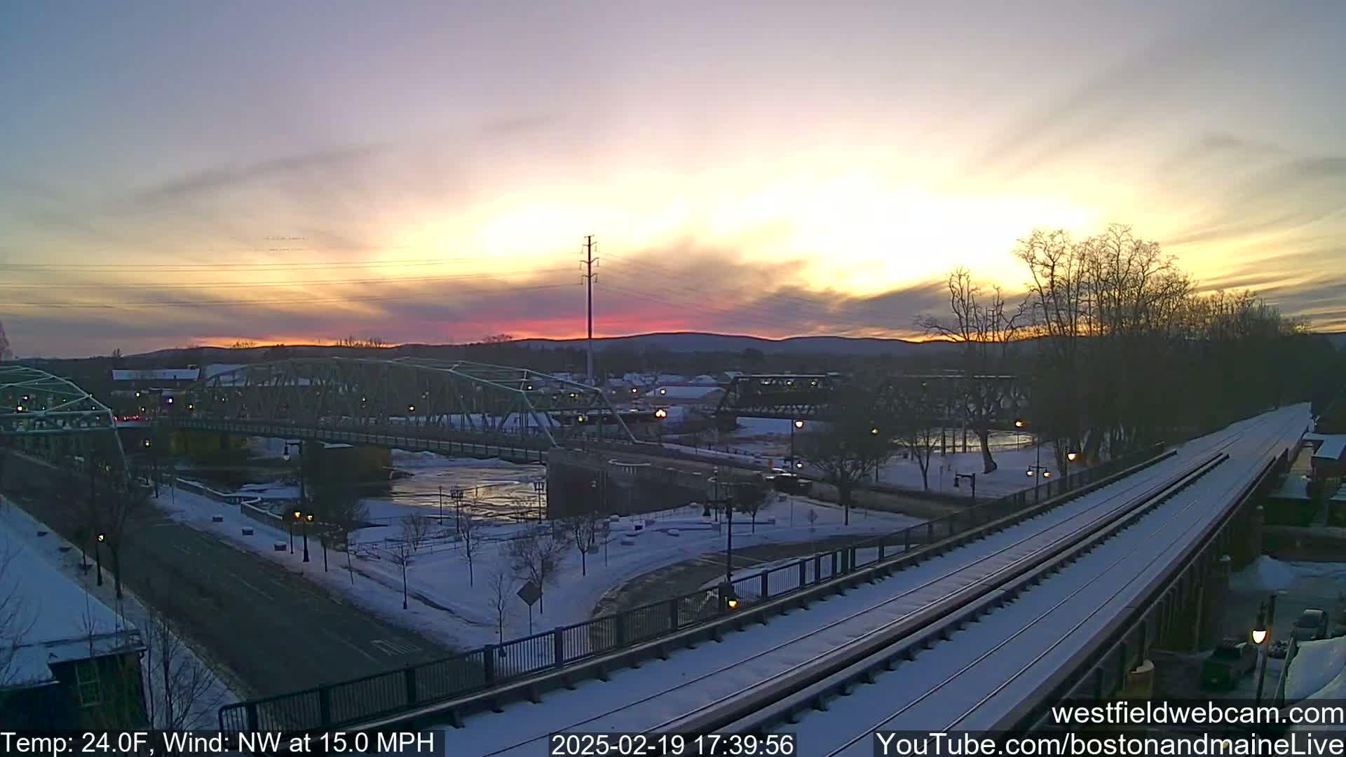A snow-covered town at sunset, featuring a river, bridges, and train tracks, with a northwest wind at 15 miles per hour and a temperature of 24 degrees Fahrenheit.