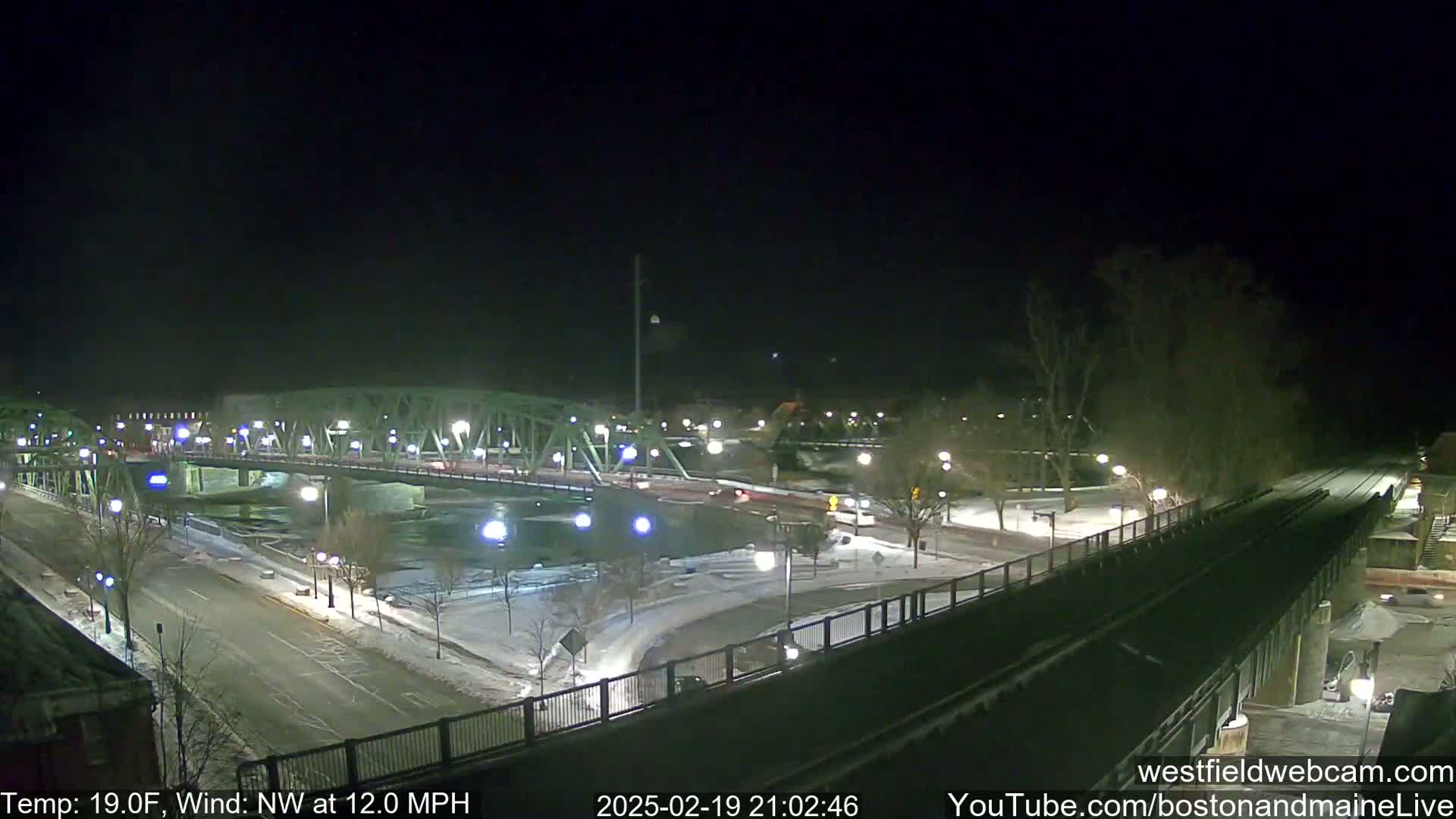 A nighttime view of a snow-covered bridge and roadway, with a river below, under a clear, dark sky with a northwest wind.