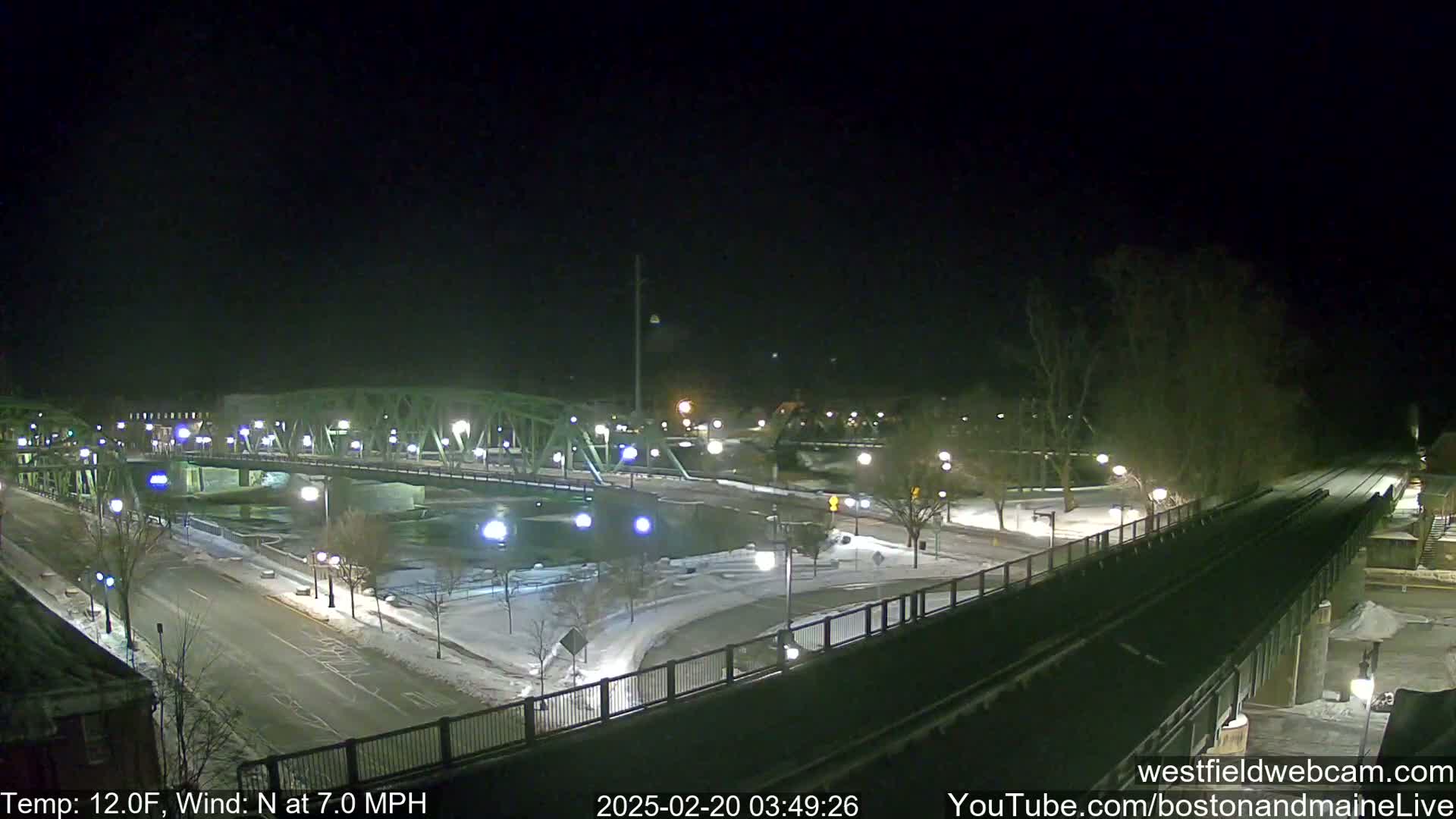 A snow-covered roadway and bridge are illuminated at night under a clear sky with a northerly wind.