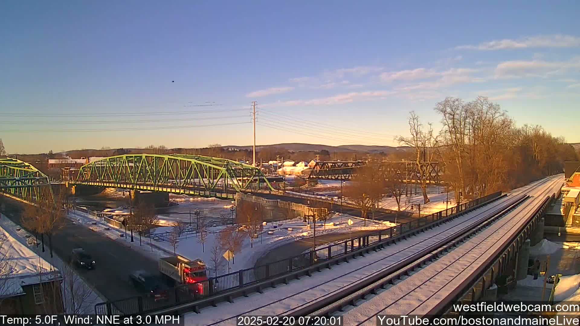 A partly sunny, cold winter morning scene shows two bridges crossing a river, with train tracks and light snow covering the ground and some vehicles on a road.