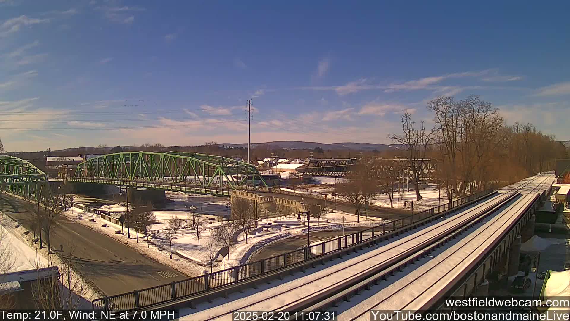 A snow-covered train track and road intersect near a green bridge over a river under a mostly clear, sunny sky.