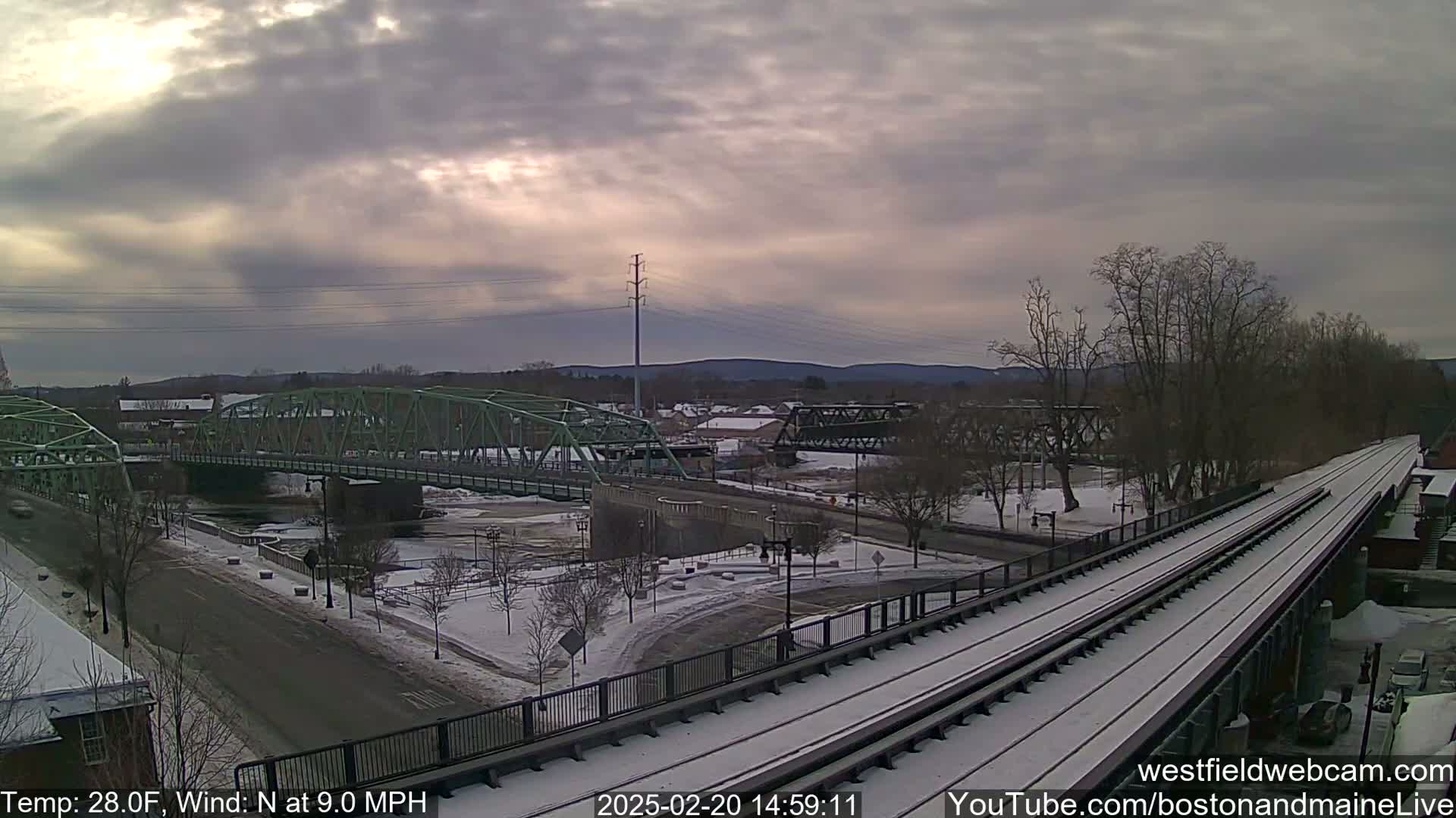 A snow-covered train track and two bridges span a river in a town under a cloudy sky.