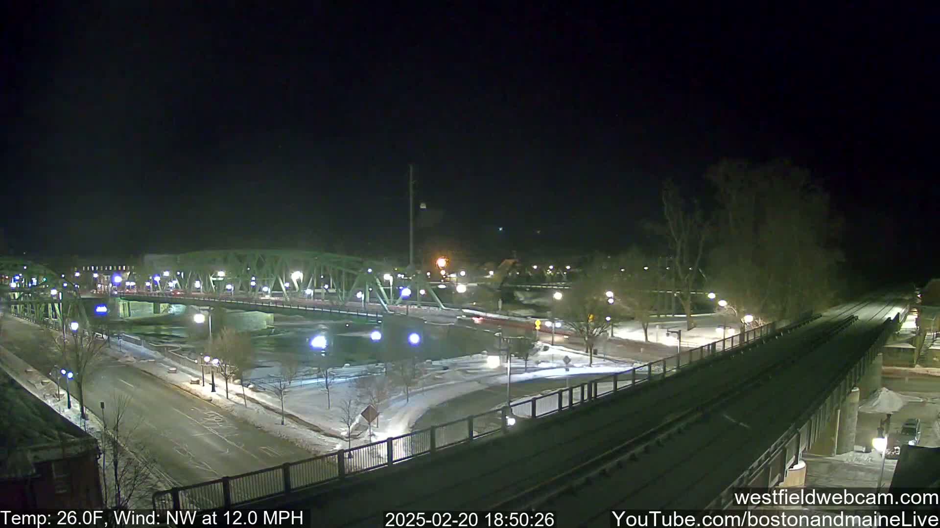 A nighttime view of a snow-covered bridge and roadway, with a river visible below, under a clear, dark sky, and a 26°F temperature with a northwest wind at 12 mph.