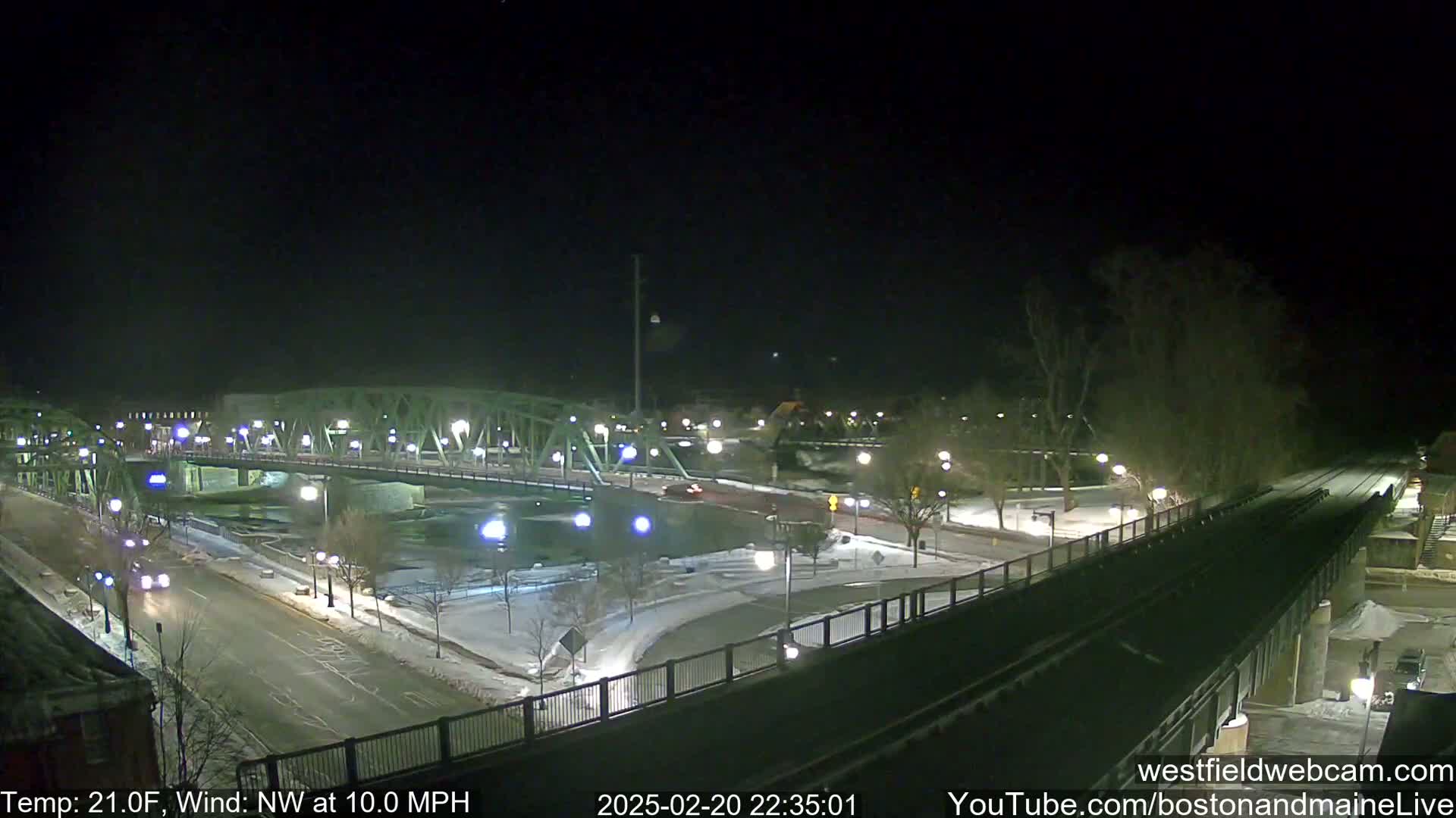 A nighttime, snow-covered scene shows a bridge over a river with sparsely trafficked roads and walkways, under a clear sky with a northwest wind of 10 mph and a temperature of 21 degrees Fahrenheit.