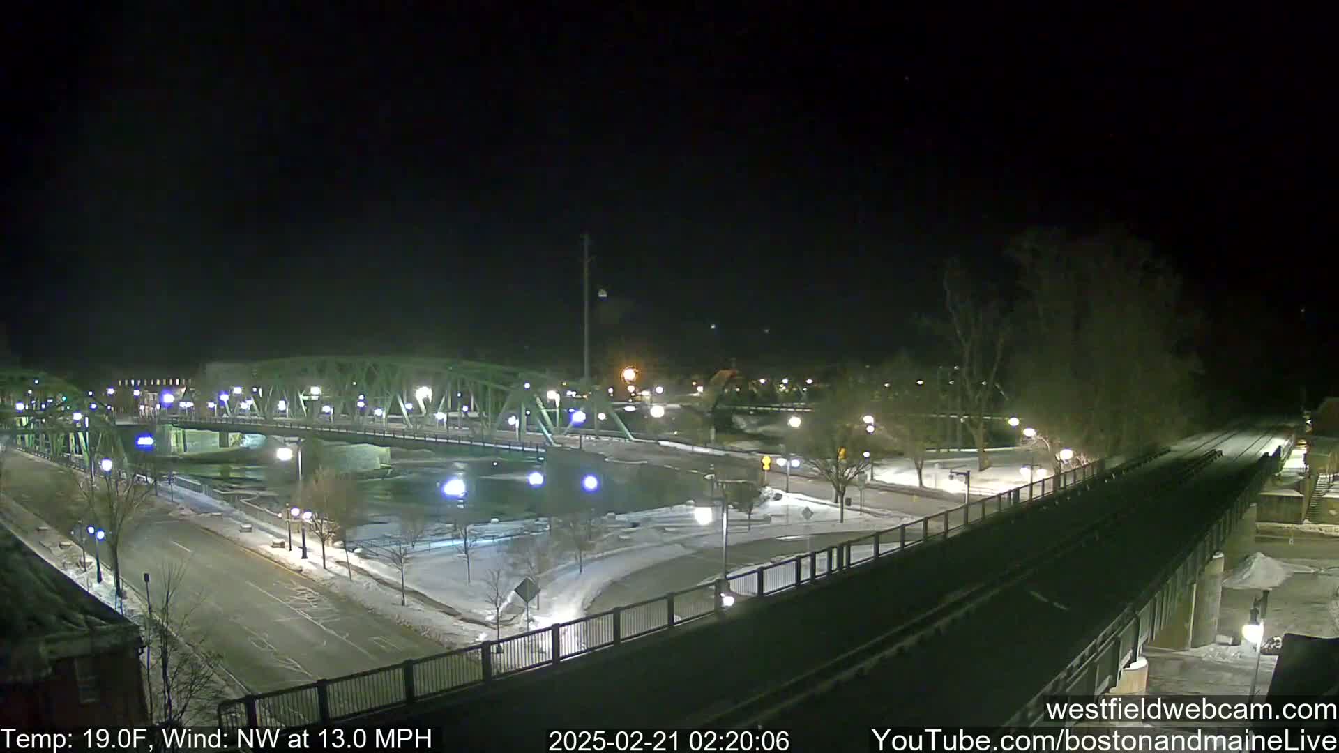 A nighttime view of a bridge over a river, with snow-covered ground and a roadway, under a clear, dark sky with a northwest wind.