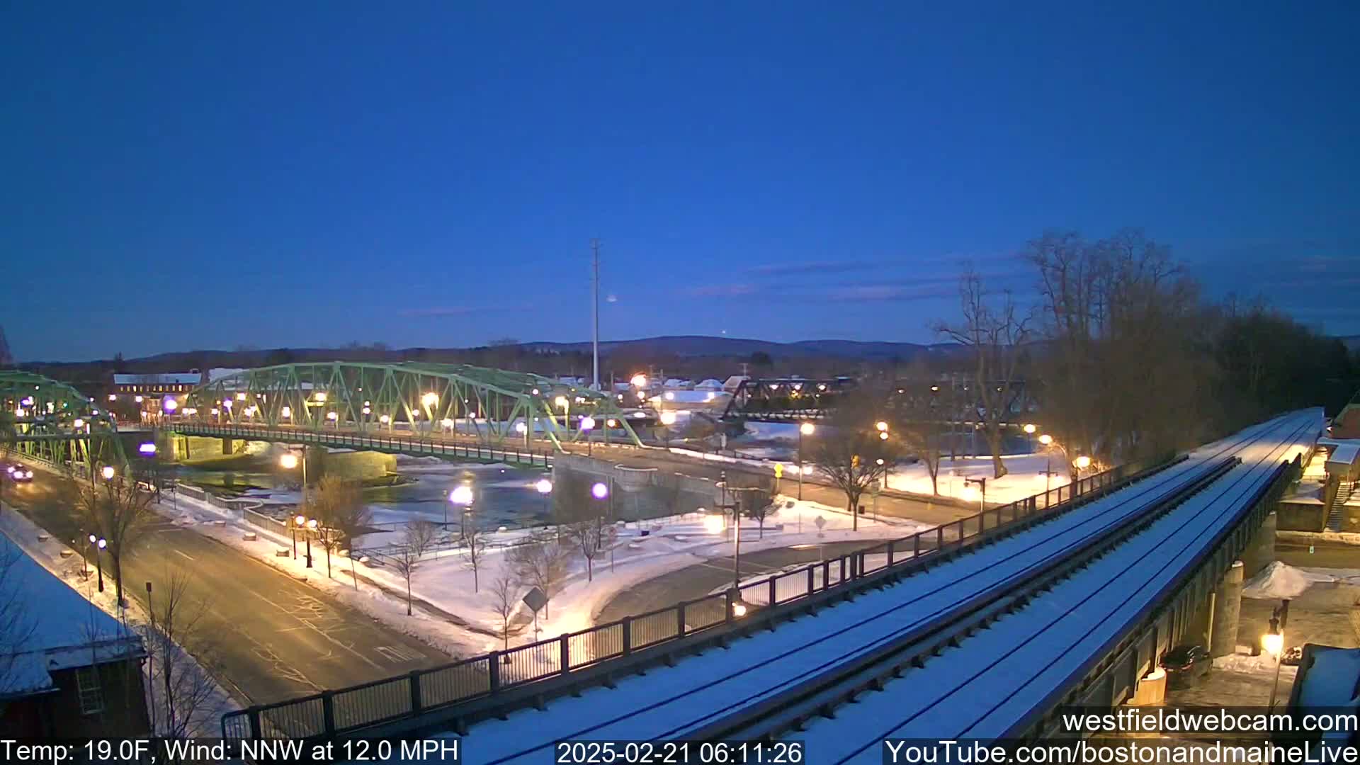 A snow-covered town scene at twilight shows a green bridge over a river, with a train track elevated alongside a road.
