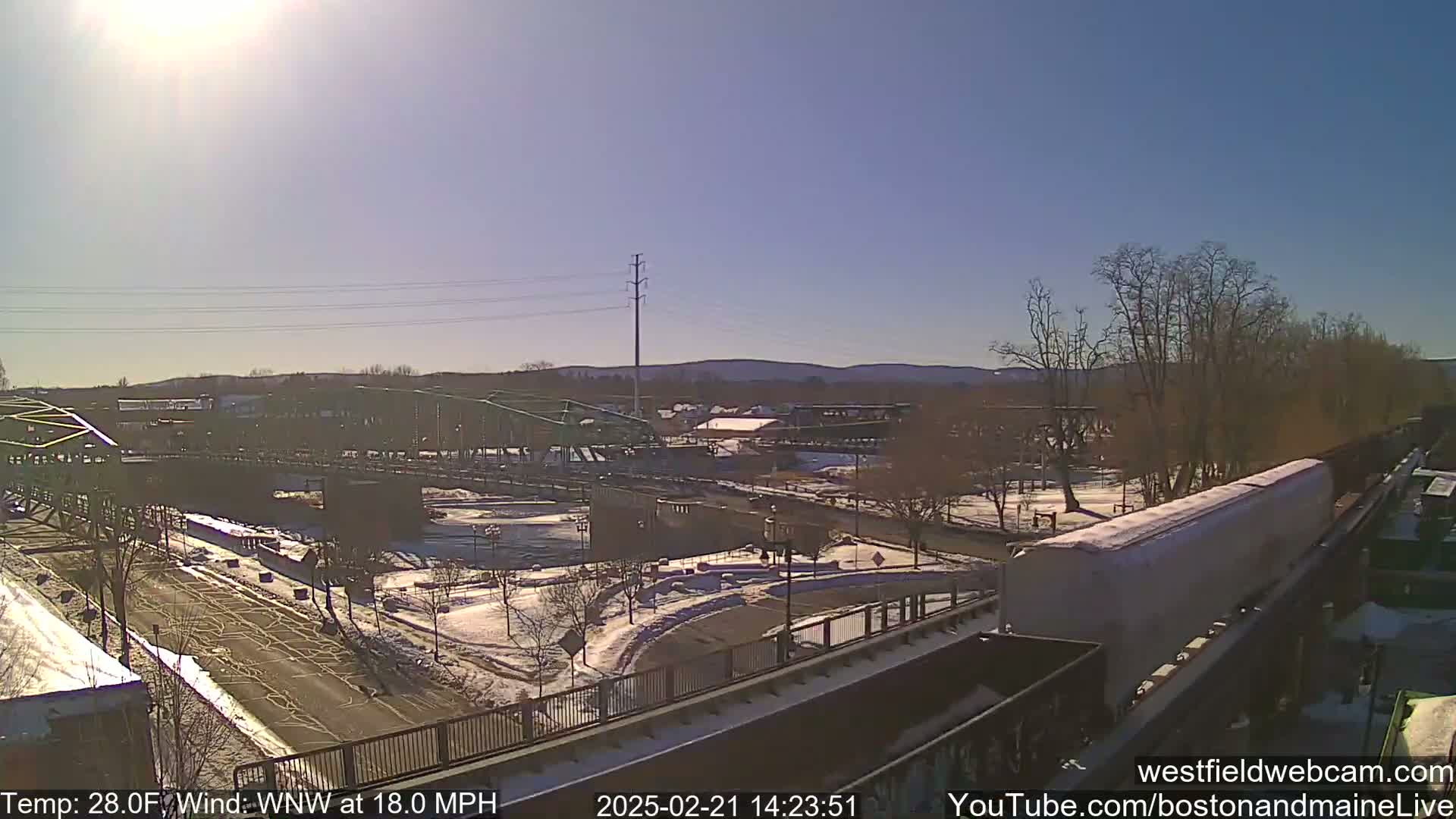 A sunny, snowy scene shows a bridge over a river, a train on tracks, and a partially snow-covered town in the distance under a bright, mostly clear sky with a WNW wind at 18.0 mph.