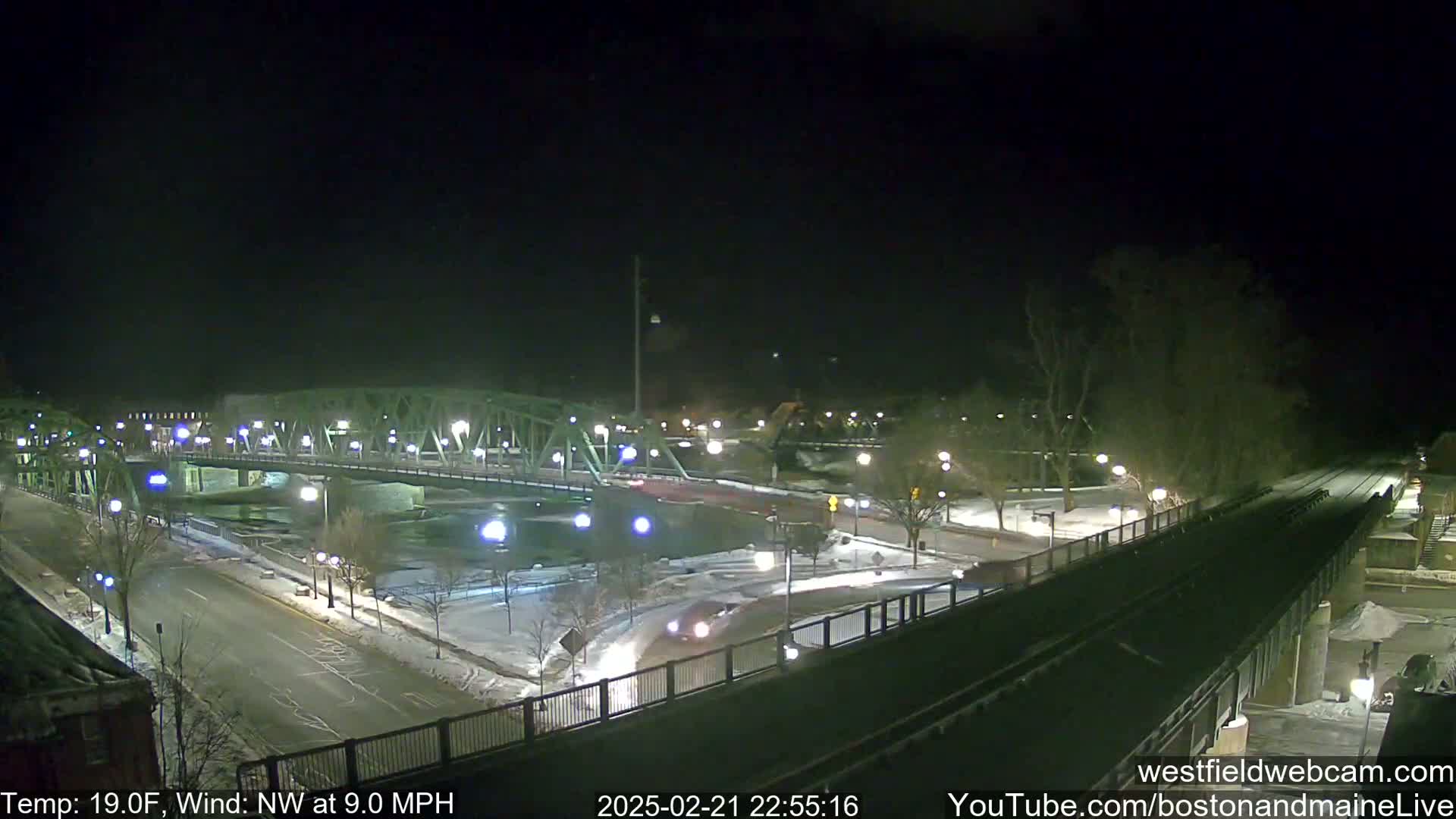 A nighttime view of a snow-covered bridge and roadway with a river below, under a clear, dark sky with a northwest wind.