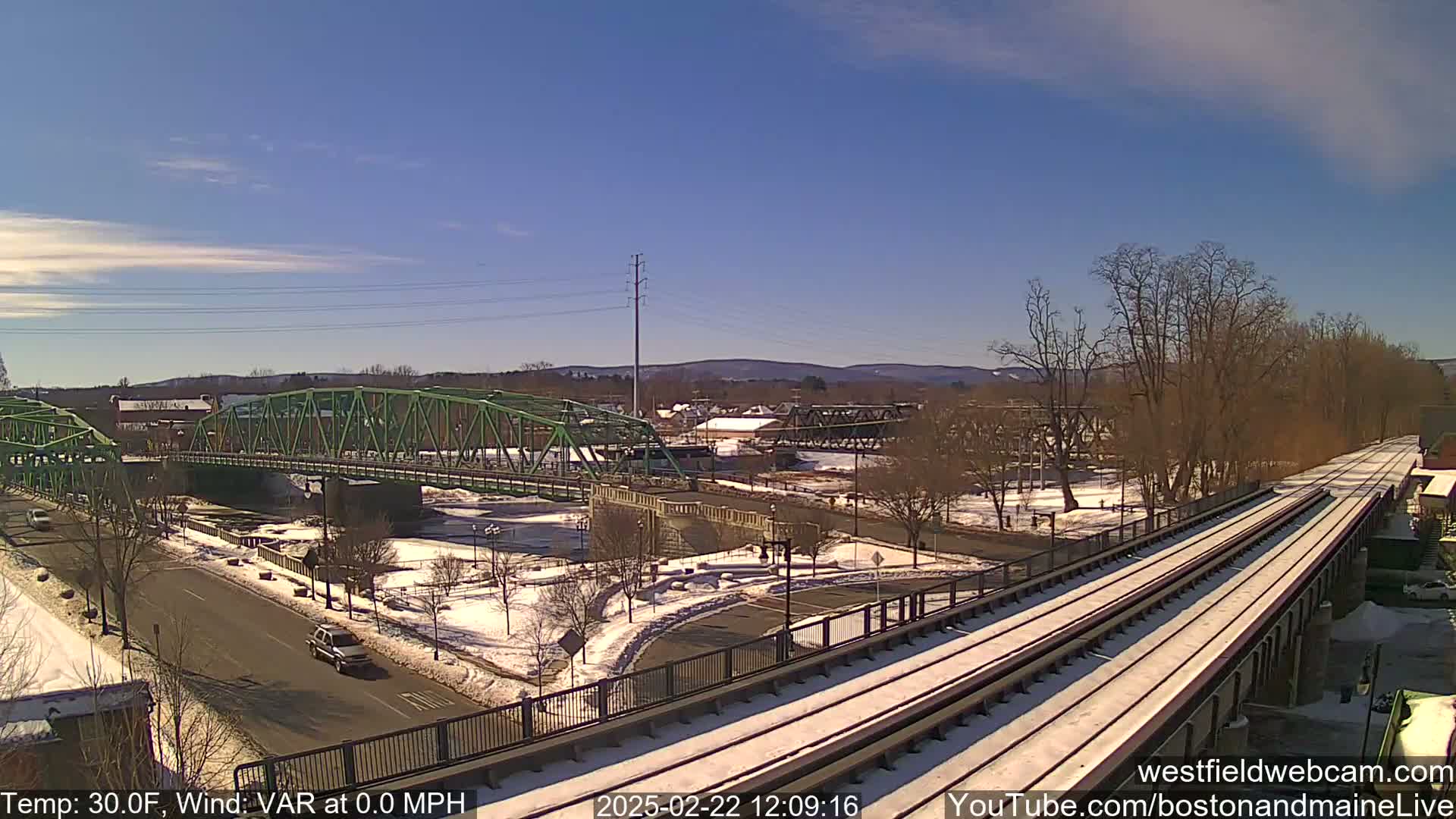 A snow-covered train track crosses a river, passing over a green bridge and a road below on a sunny day.